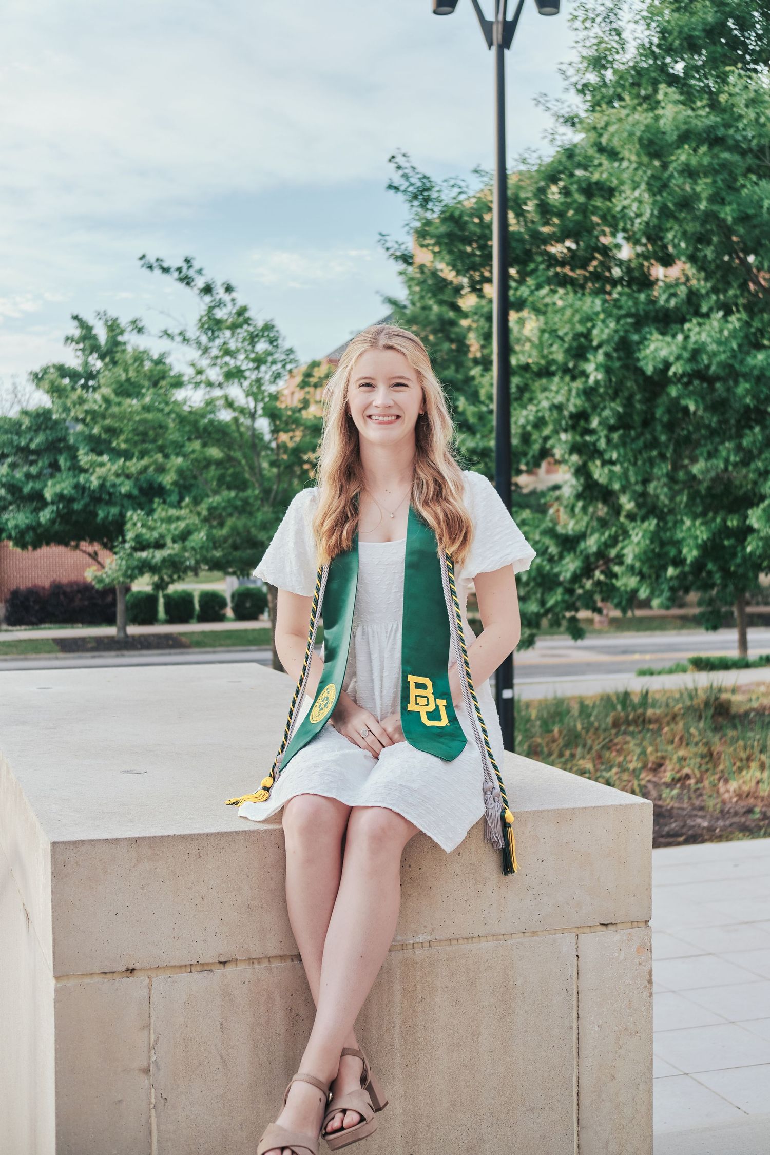 Baylor University graduate in white dress and green BU sash sitting on a stone ledge on campus.