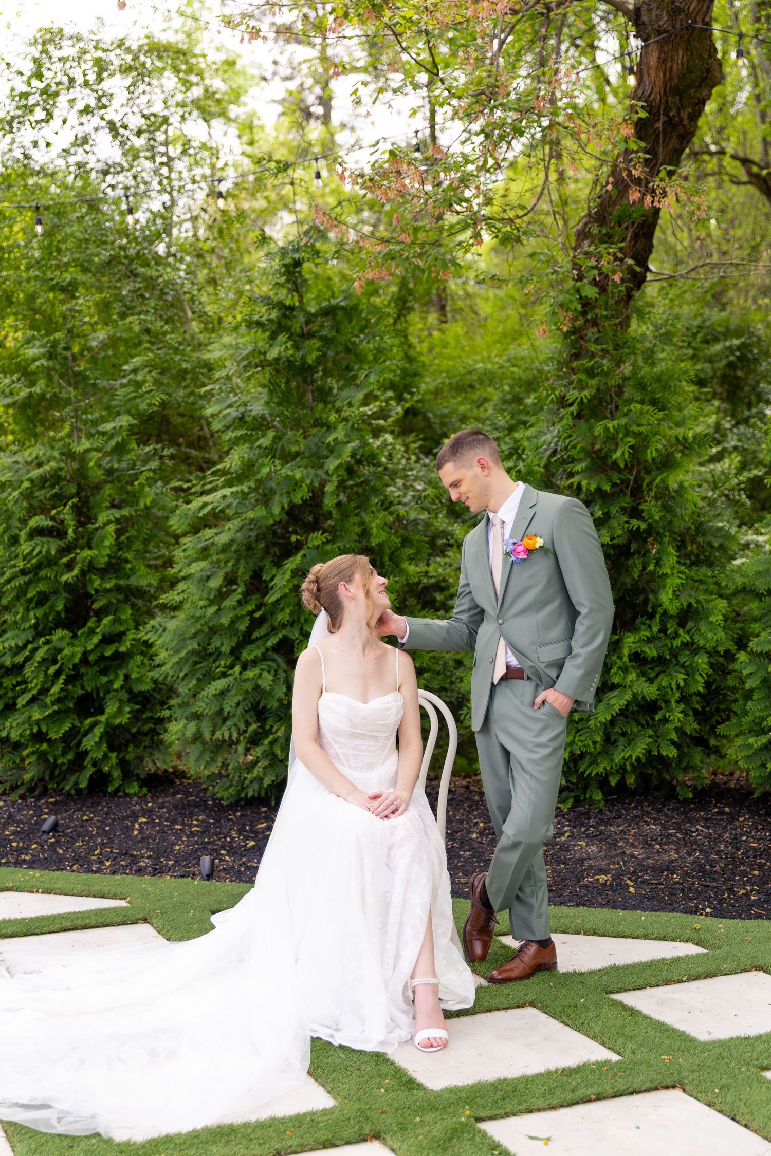 Groom in sage green suit tenderly touches brides hair as she sits in a chair at an outdoor garden wedding.