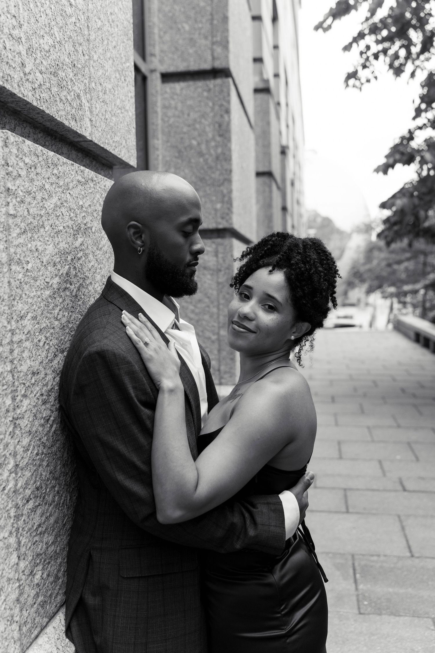Black and white portrait of a Black couple embracing tenderly against a granite wall on a city street.