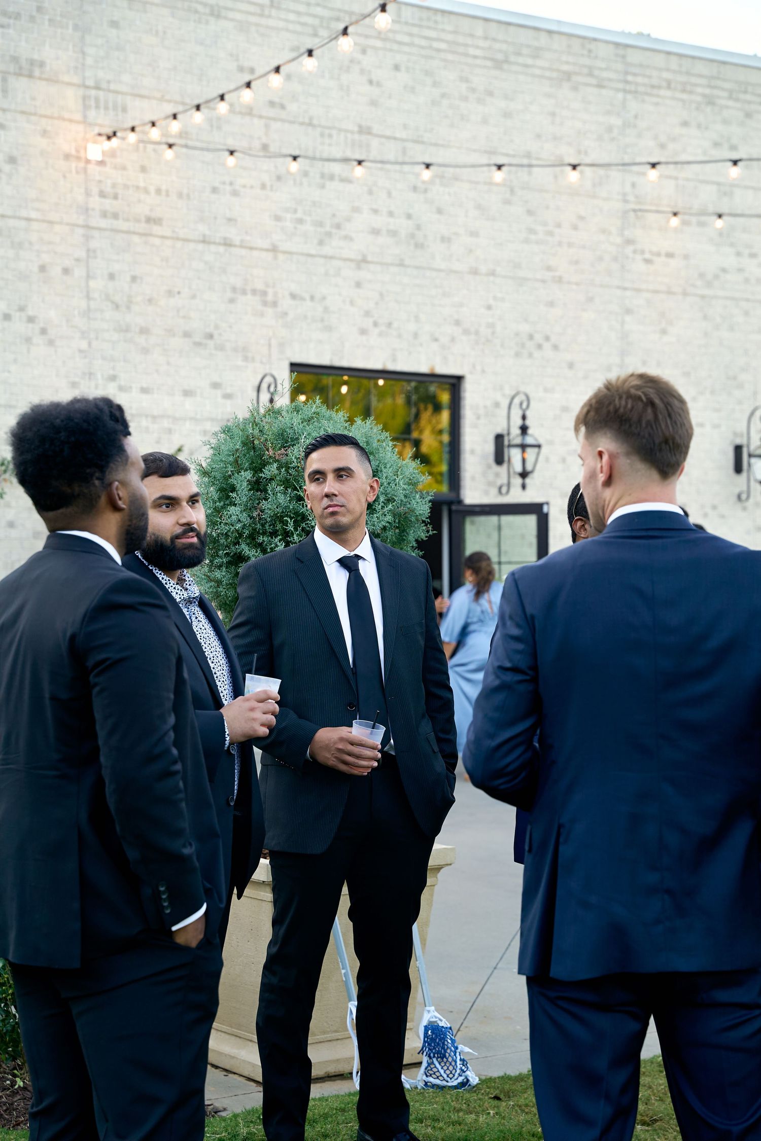 Formally dressed groomsmen in black suits socialize outdoors at an evening wedding reception.