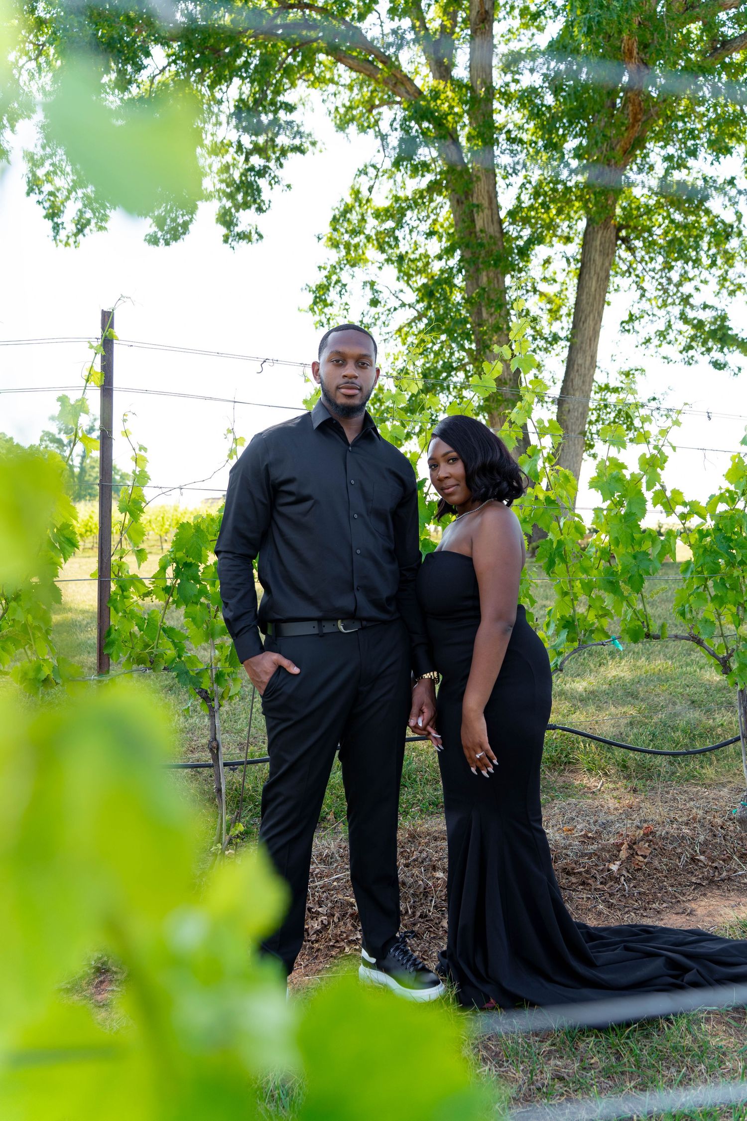 Stylish couple poses together in all-black outfits surrounded by green vineyard foliage.