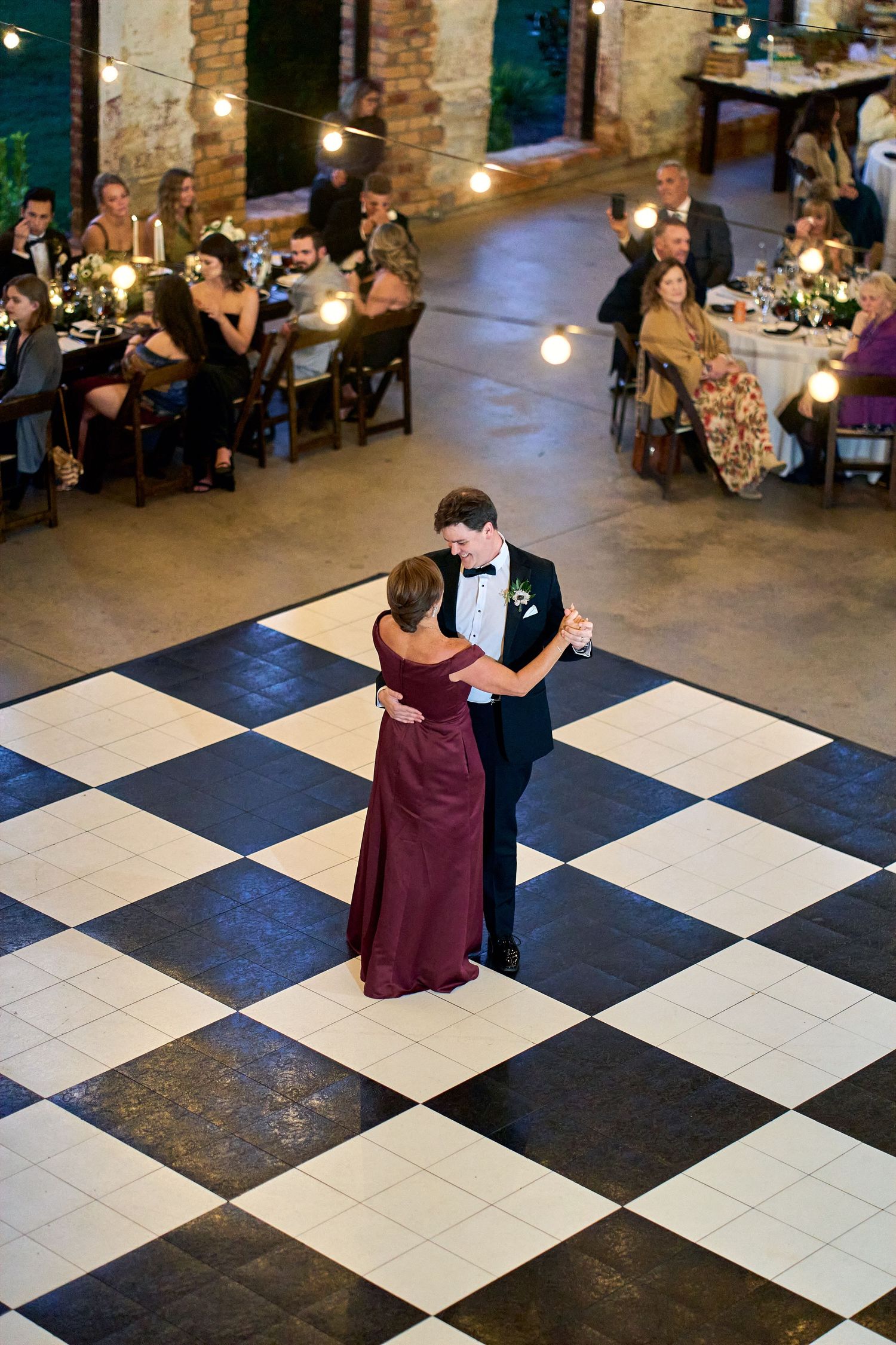 A couple shares a dance on a checkered ballroom floor while wedding guests watch from tables around the perimeter.