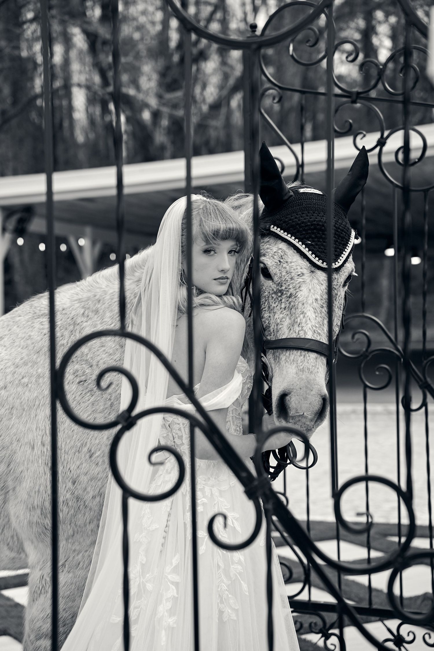 Black and white photo of a bride in a flowing dress posing with a white horse near ornate wrought iron gates.
