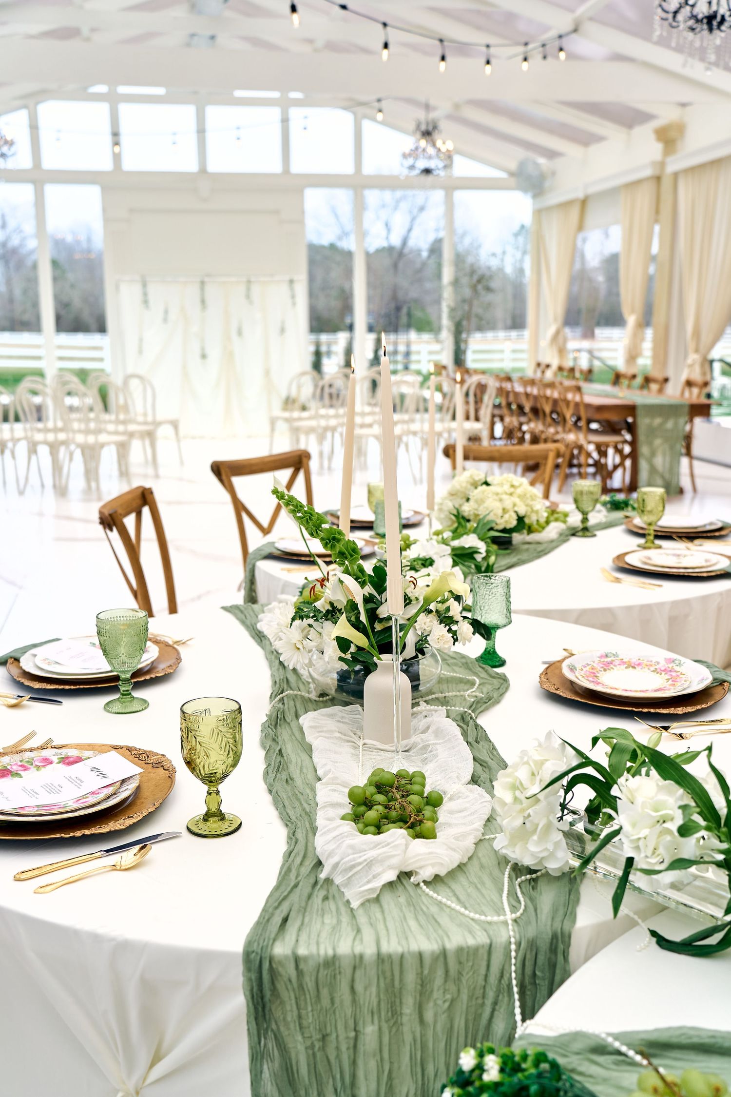 Elegant wedding reception table with white linens, green chiffon runners, and floral centerpieces in a bright sunlit venue.
