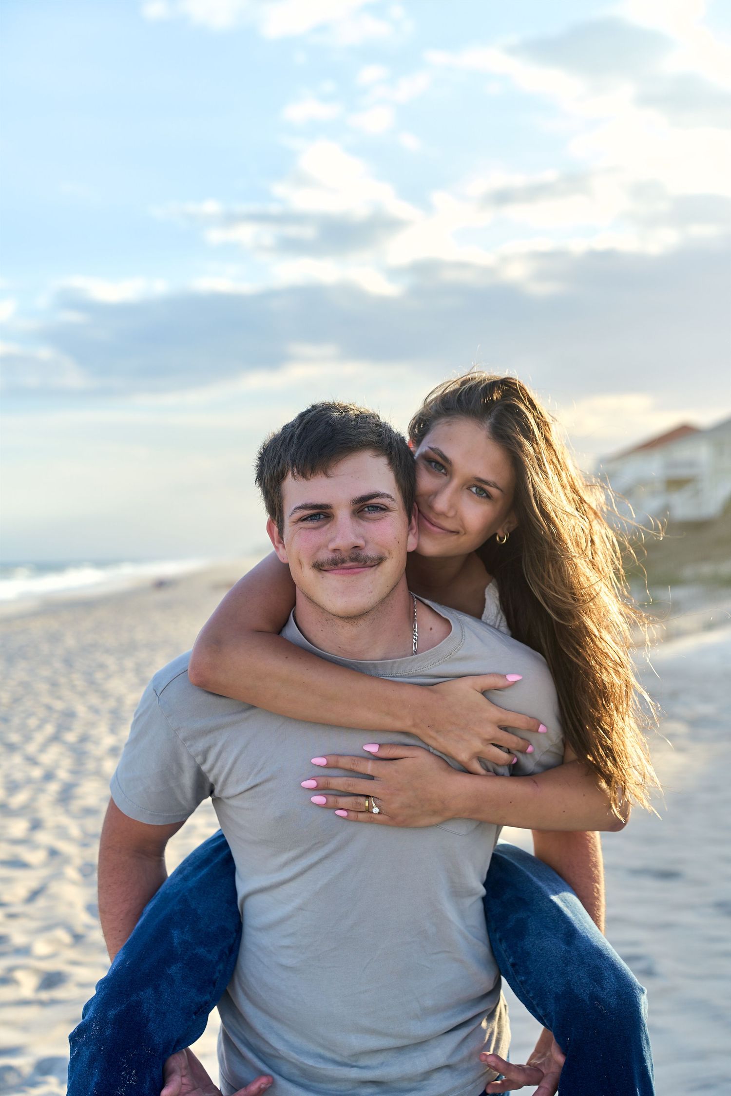 Two people sharing a playful piggyback ride moment together on a sunny beach with ocean waves in the background.