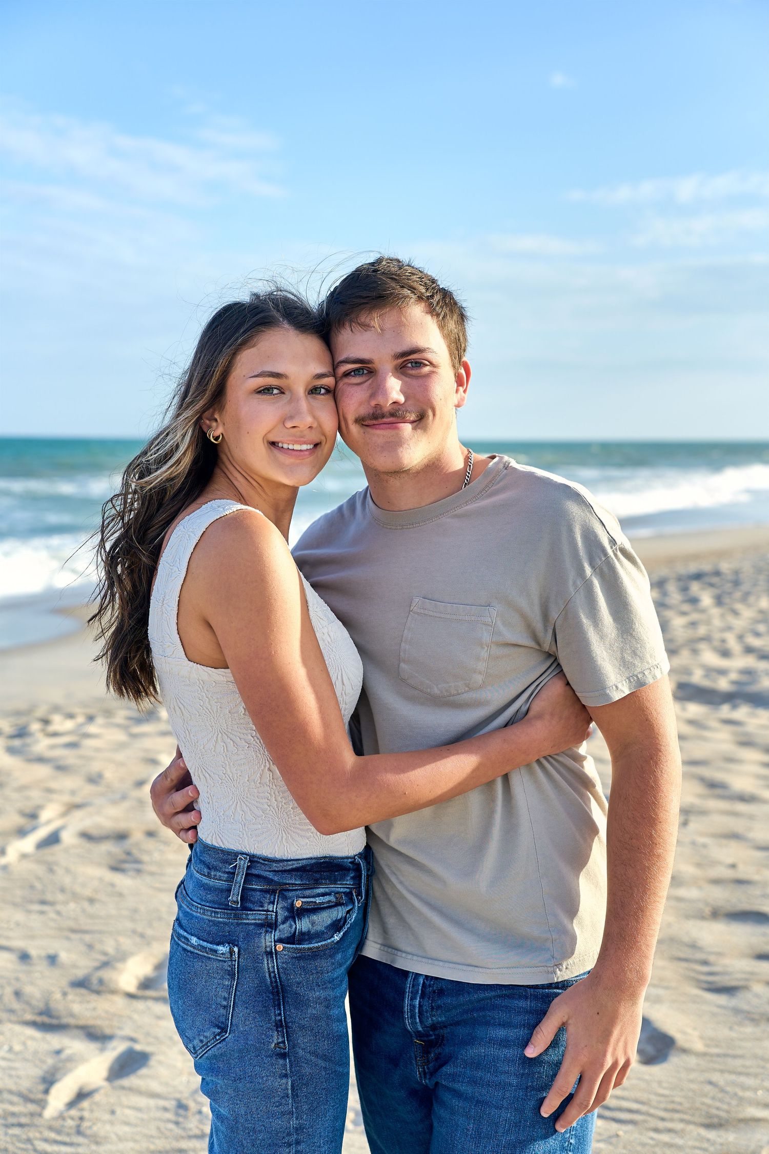A couple shares a tender moment together on a sunny beach with ocean waves and blue sky in the background.