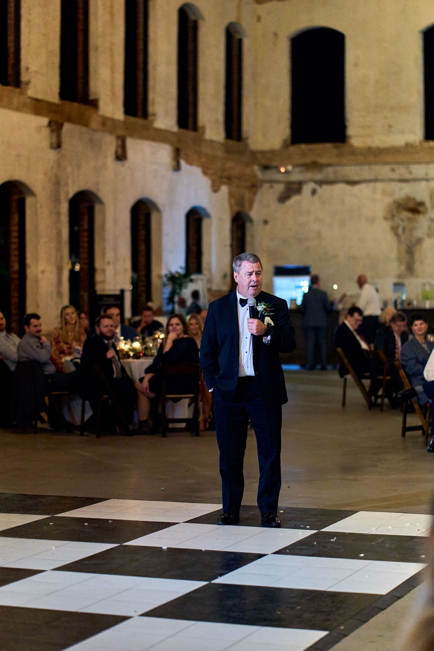 Someone gives a speech standing on a black and white checkered dance floor at an elegant indoor reception venue.