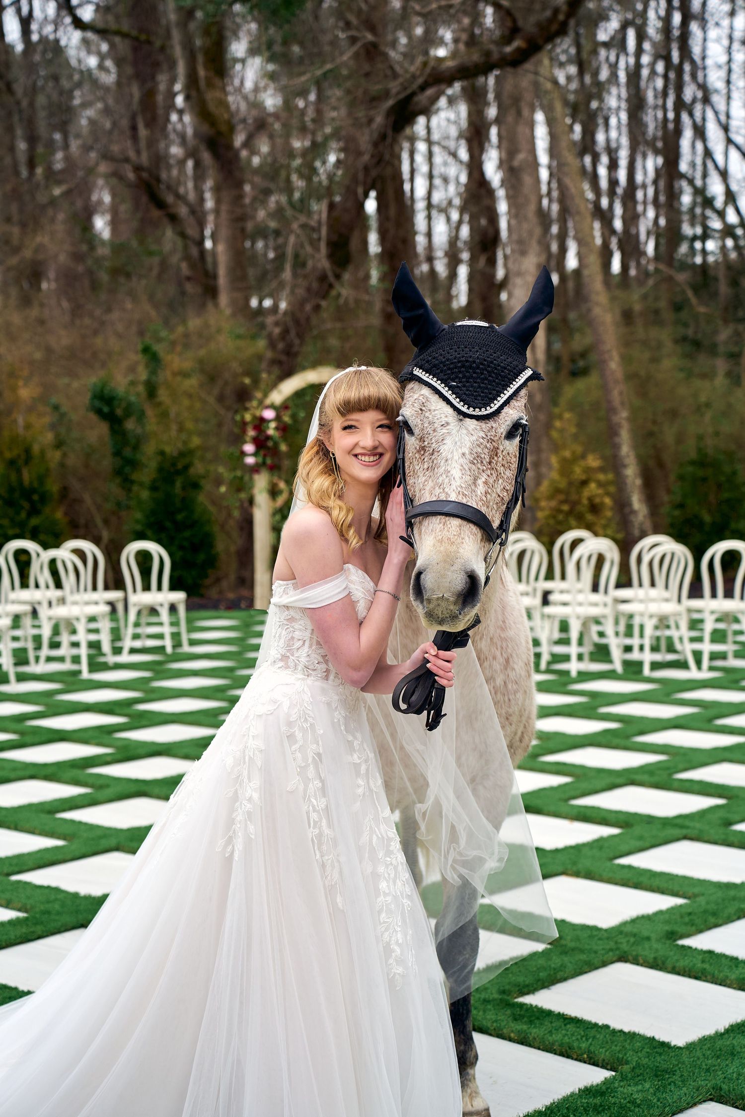 A person in a white wedding dress poses with a decorated white horse on a checkerboard grass lawn with white chairs.