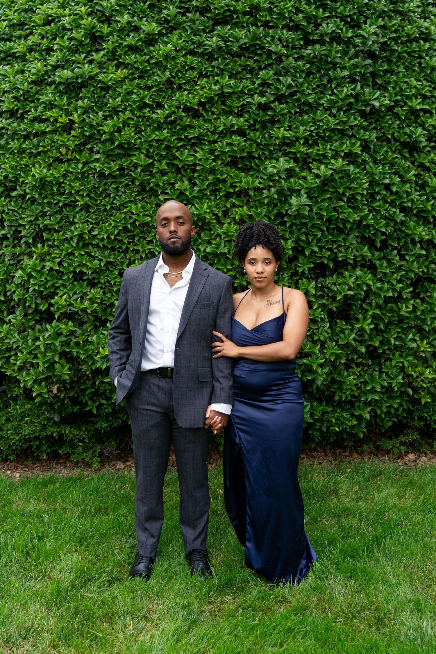 An elegant couple in formal attire poses seriously in front of a lush green hedge wall.