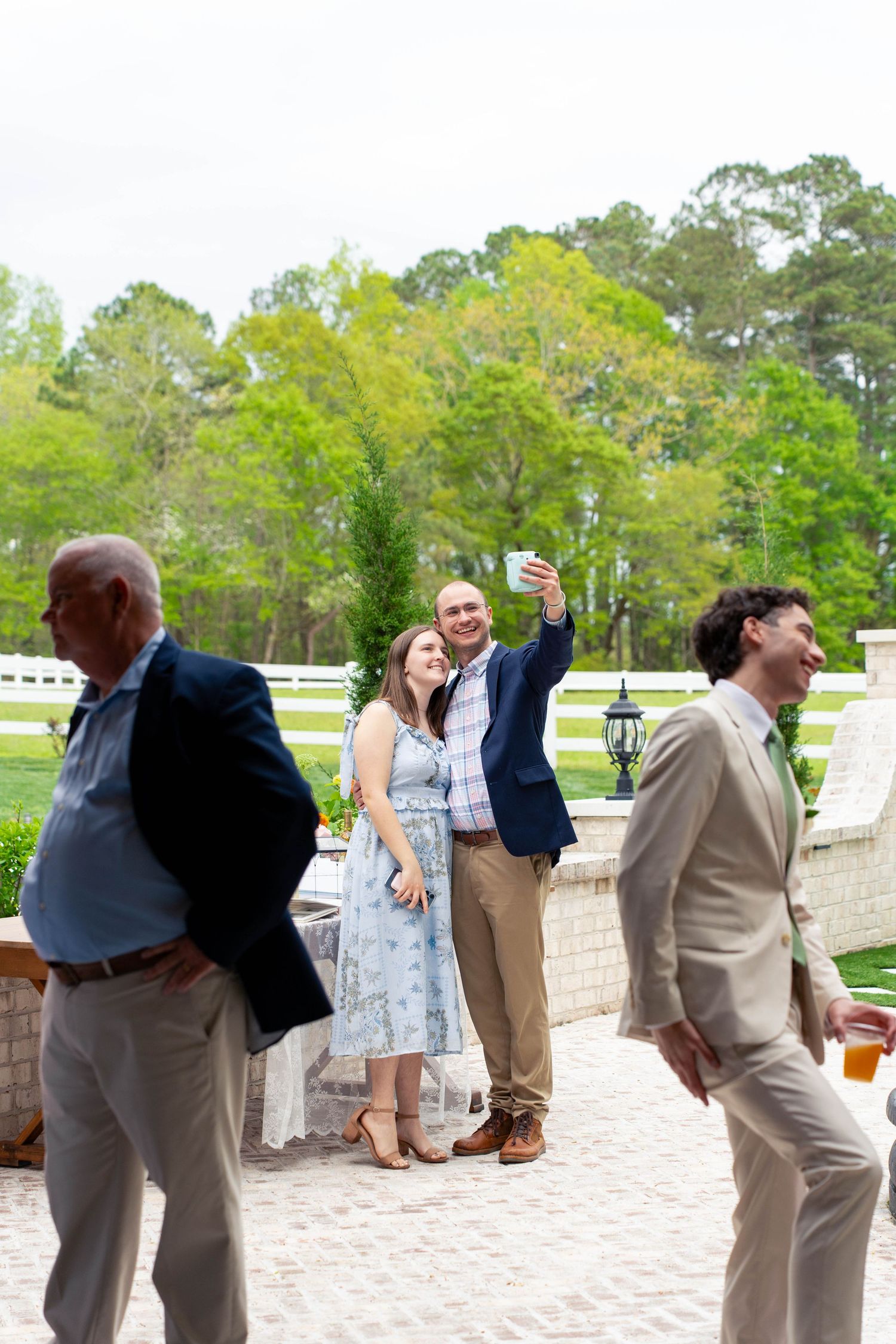 Wedding guests taking a selfie outdoors at an elegant venue with white fencing and lush green trees.