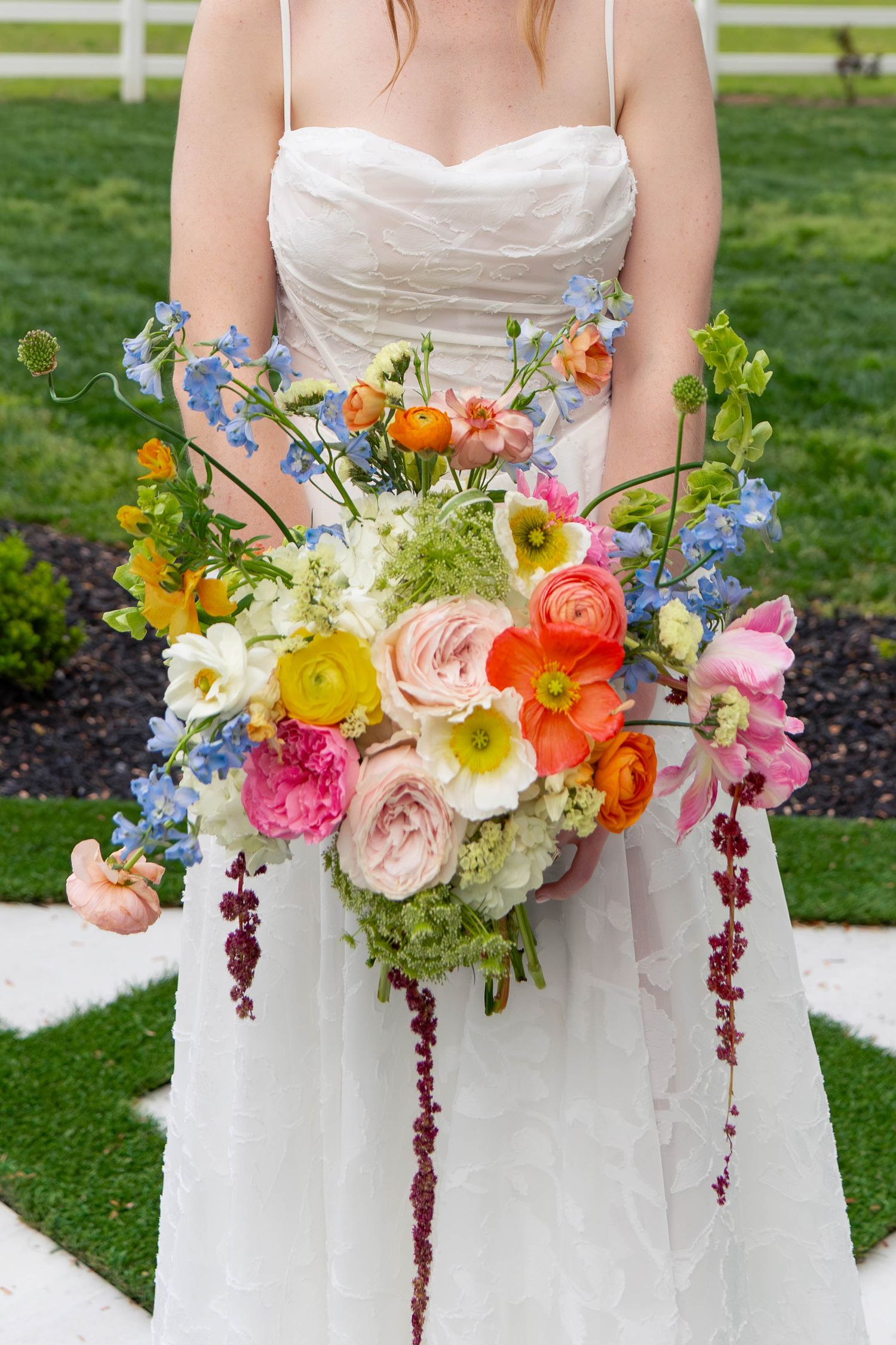 Bride holds a vibrant colorful wedding bouquet with ranunculus, poppies, and blue delphinium in white lace gown.