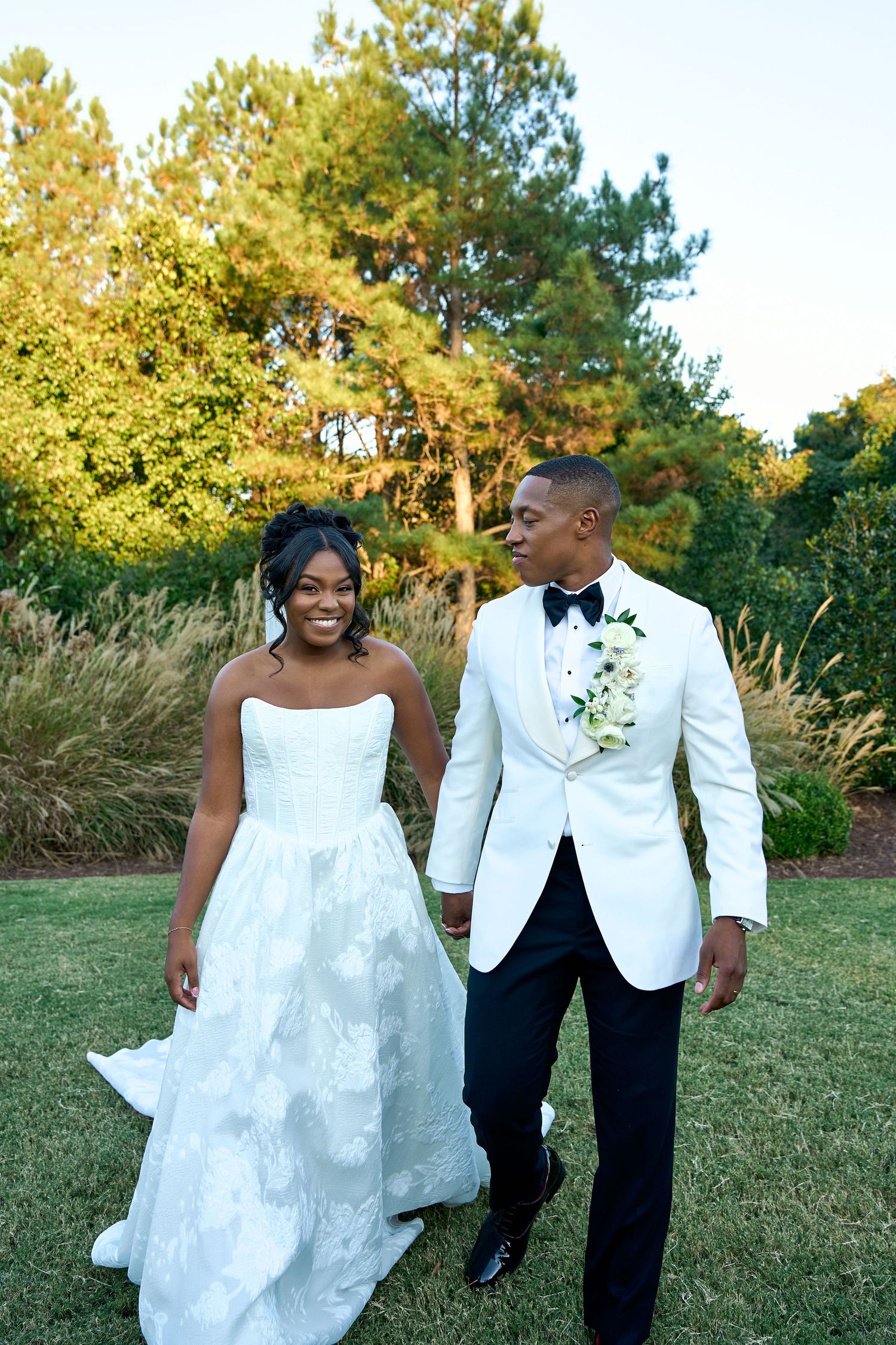 Newlywed couple walks hand in hand outdoors, groom in white tuxedo jacket and bride in strapless gown.