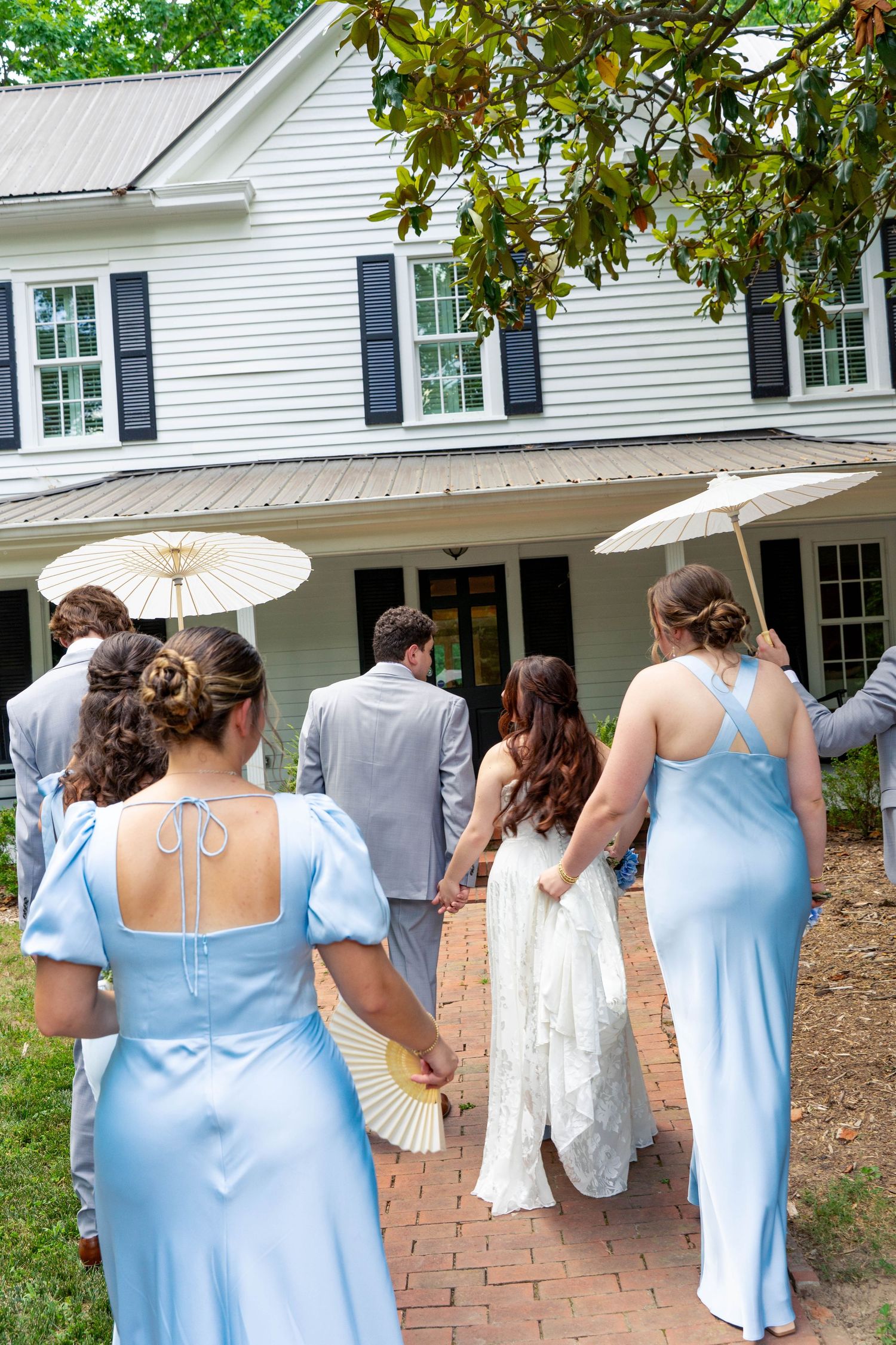 Wedding party walks toward a white colonial house, bridesmaids in light blue dresses carrying parasols and fans.