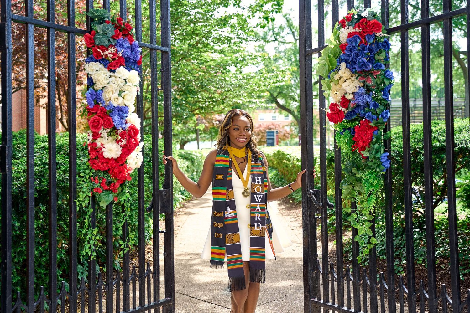 Graduate celebrating in colorful stole walking through decorated iron gates with red, white, and blue flowers.