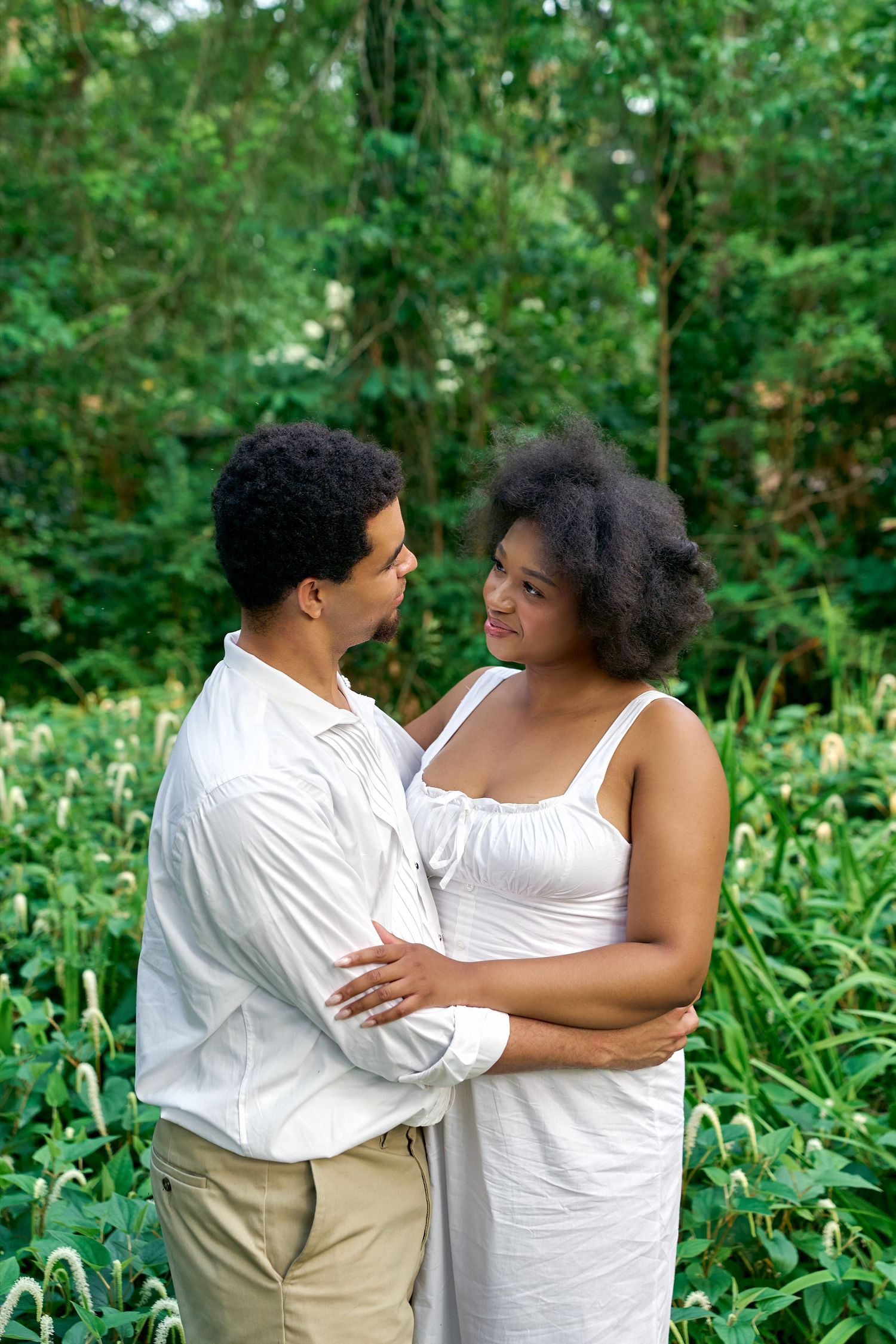 A couple in white outfits share tender moments together while posing in a lush green garden setting.
