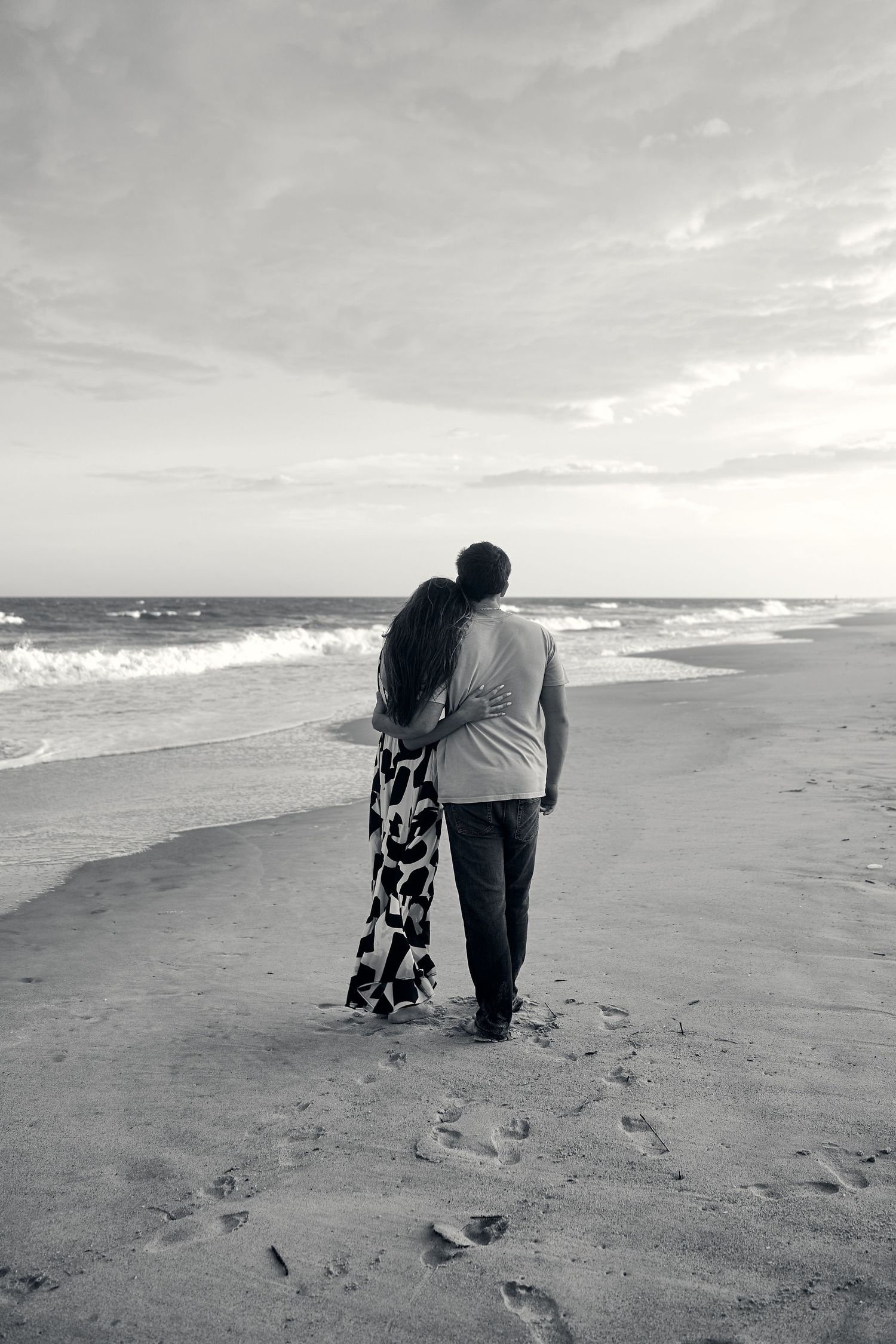 Black and white photo of a couple walking along a beach shoreline with ocean waves rolling in.