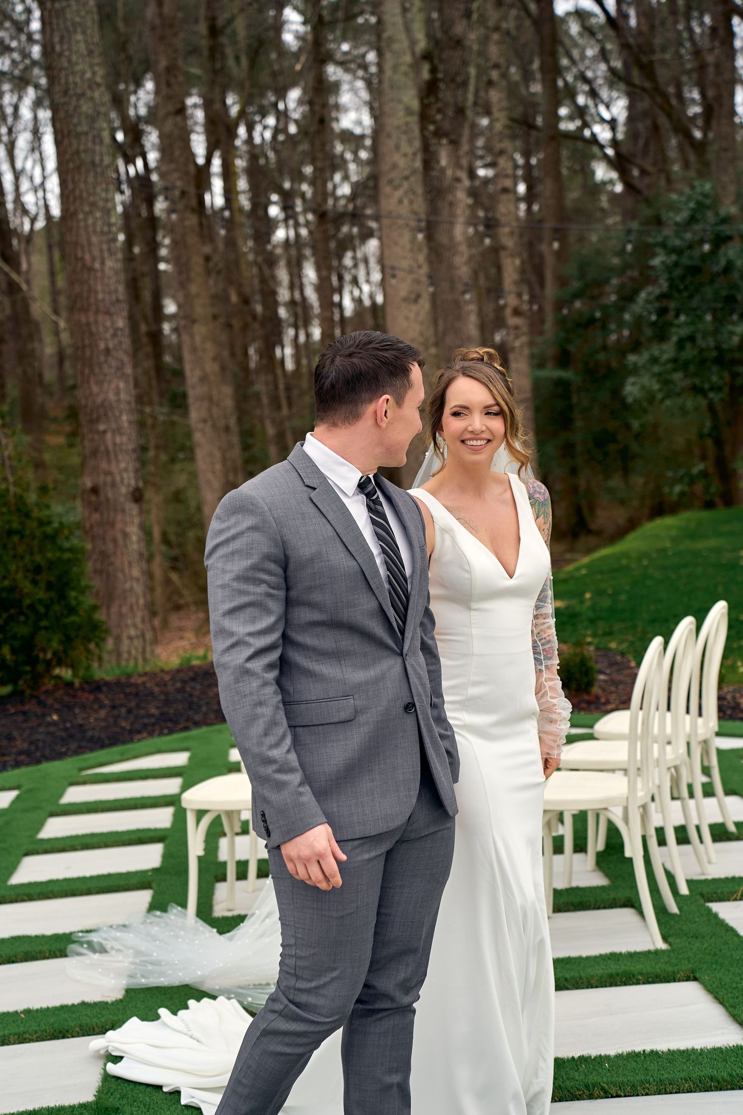 A couple shares a joyful moment during their wedding ceremony on a path lined with forest trees and white chairs.