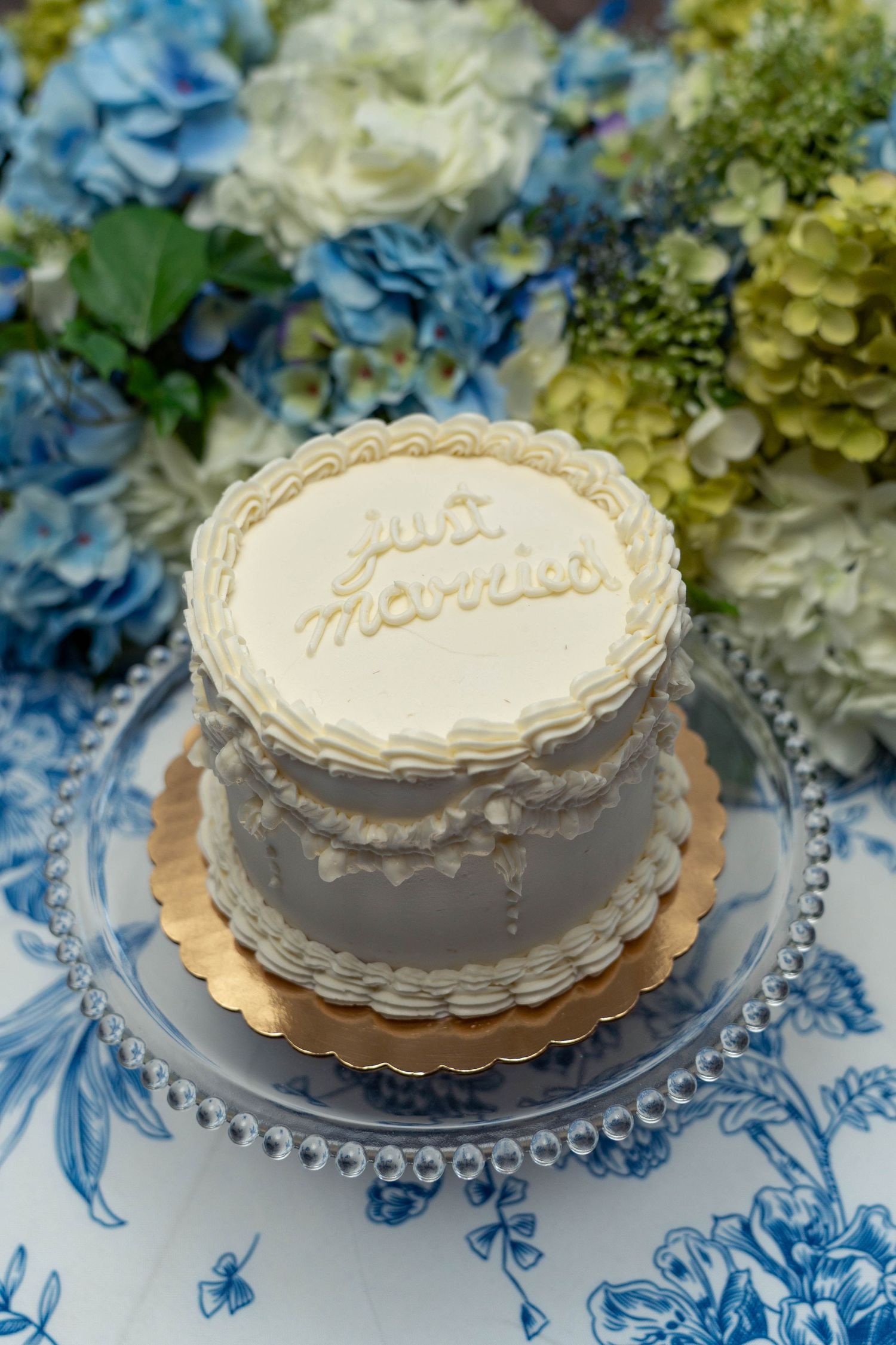 Small white wedding cake reading just married on glass stand with blue hydrangeas.