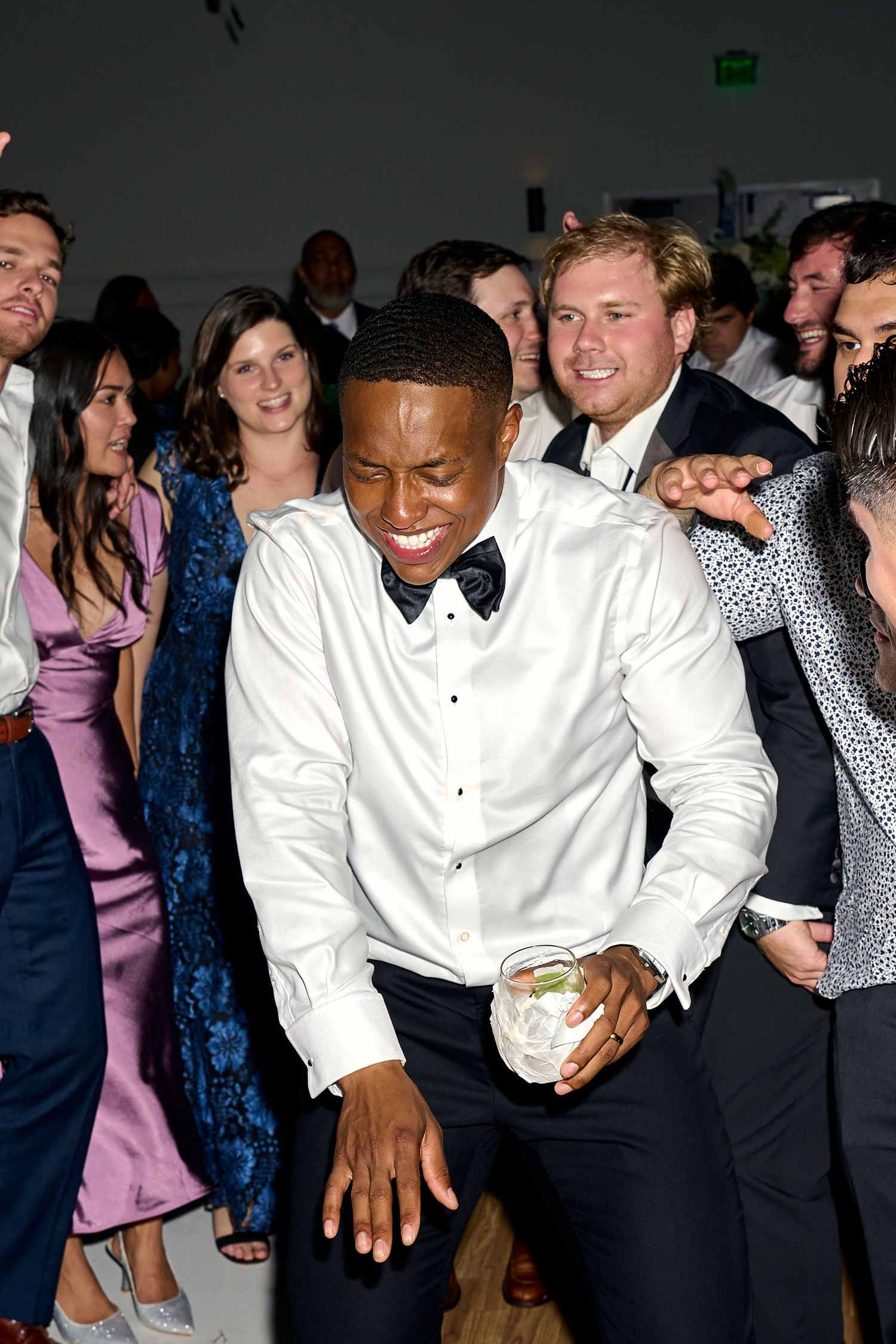 Smiling wedding guest in white shirt dances energetically holding a drink at a lively reception.