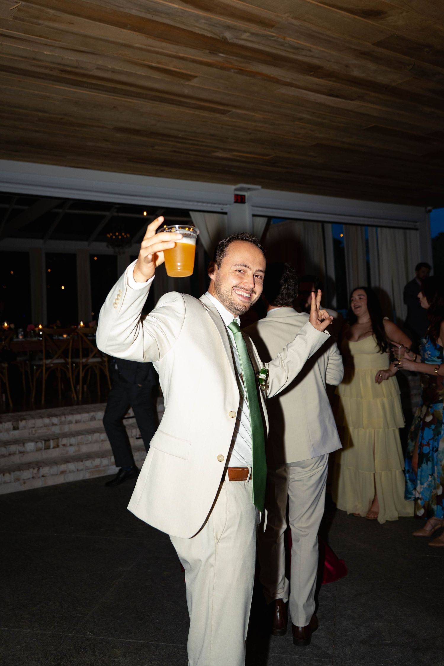 Smiling wedding guest in white suit raises beer glass while dancing at lively wedding reception.