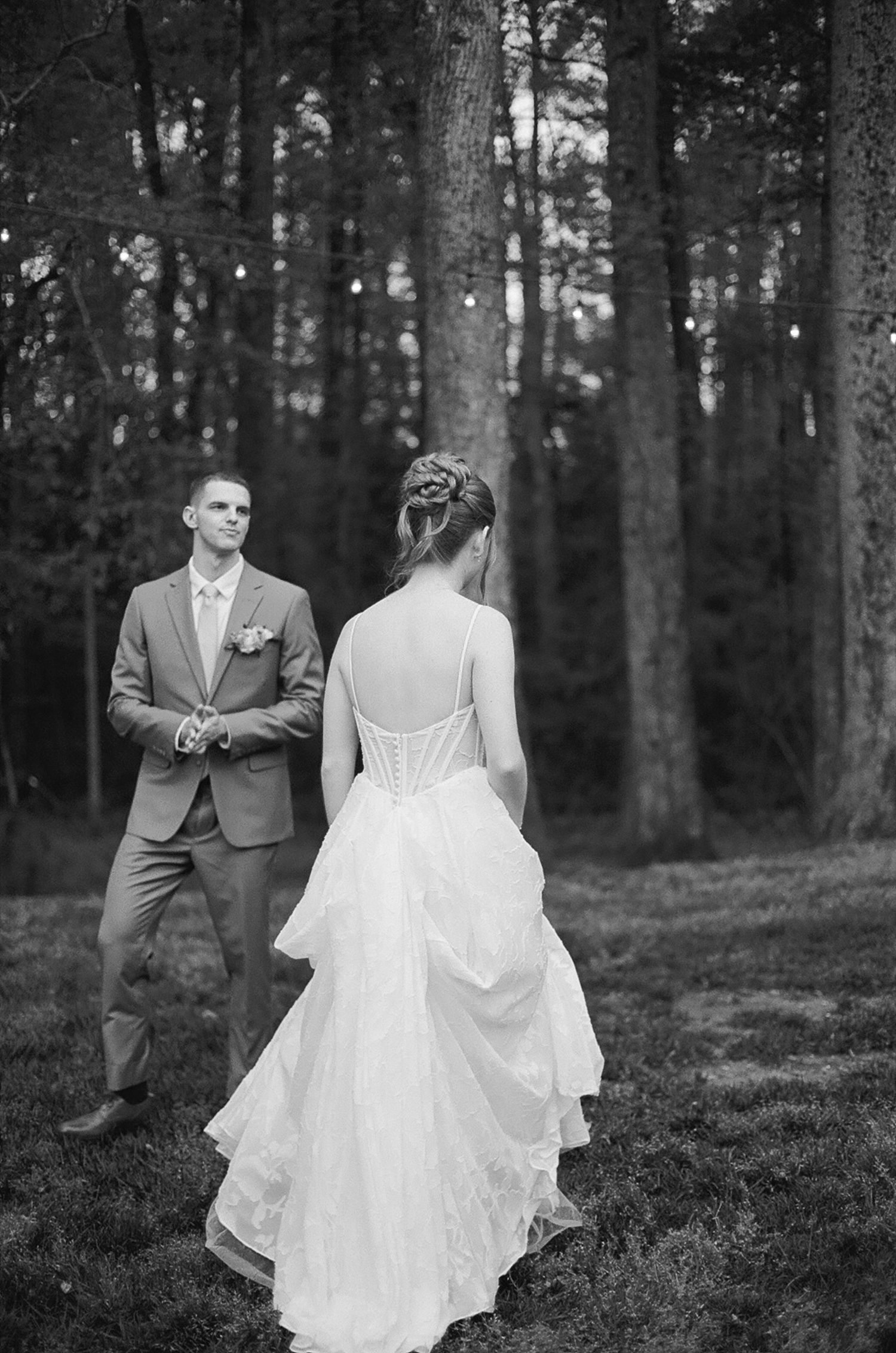 Black and white photo of bride in ruffled gown approaching groom in pine forest.