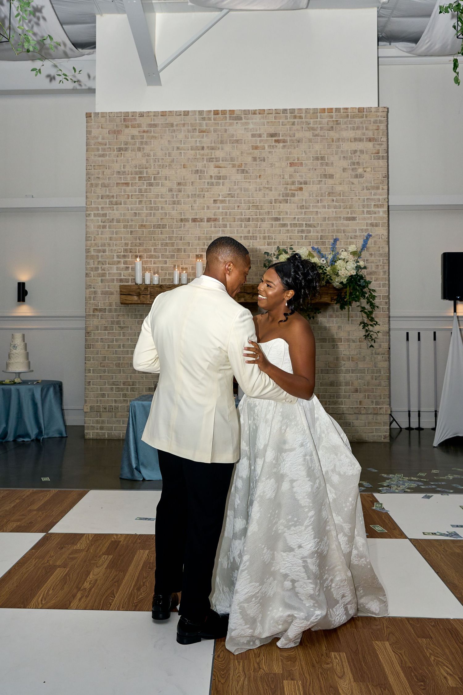 Bride in floral ball gown and groom in white jacket share first dance before brick wall backdrop.