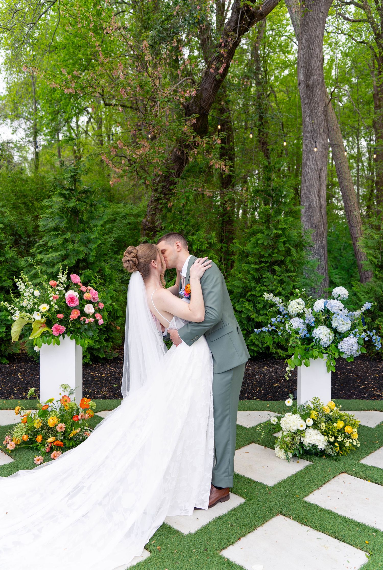 Bride and groom share first kiss at altar surrounded by vibrant floral arrangements in garden setting.