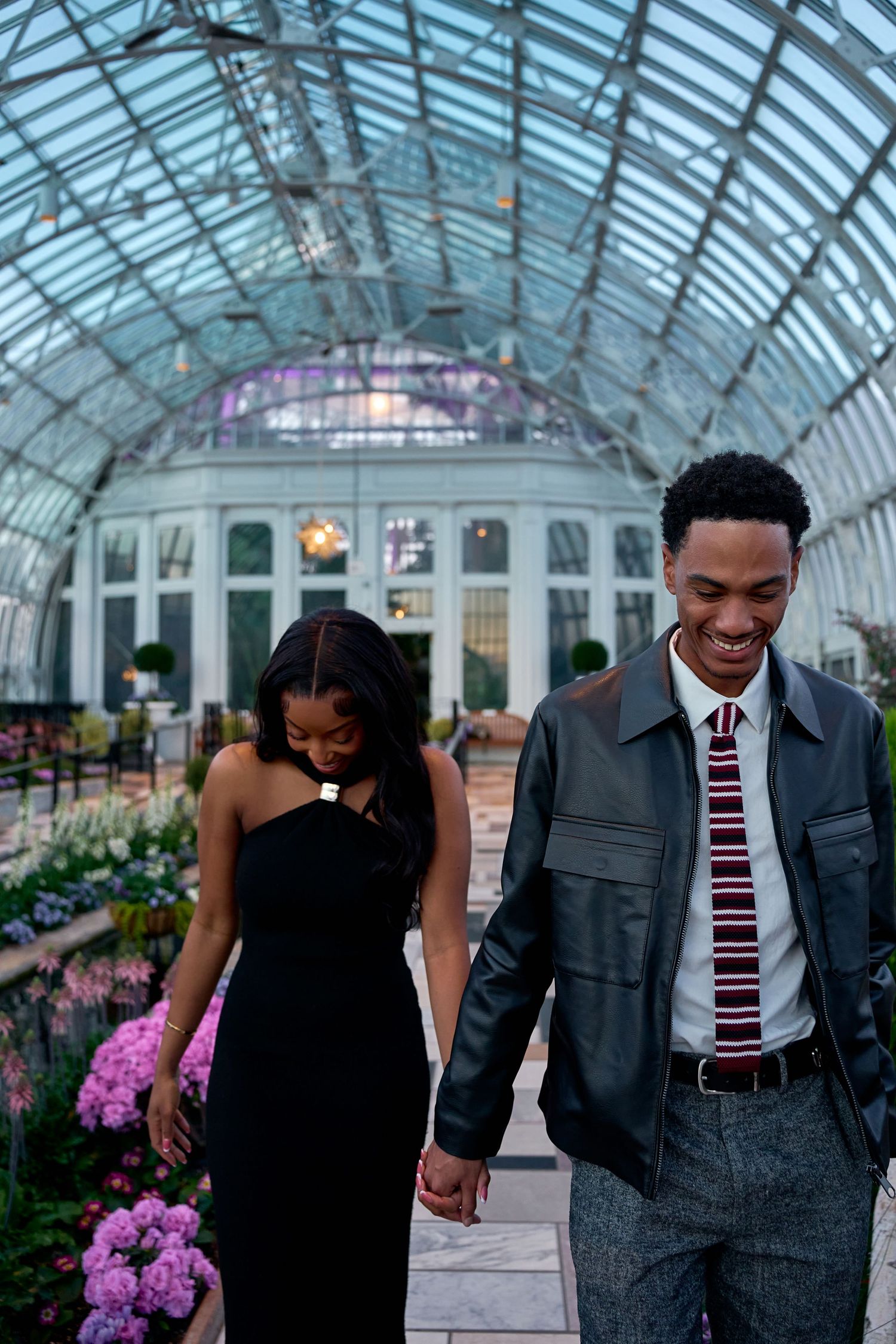 Couple holding hands and smiling while walking through a glass-domed conservatory filled with flowers.