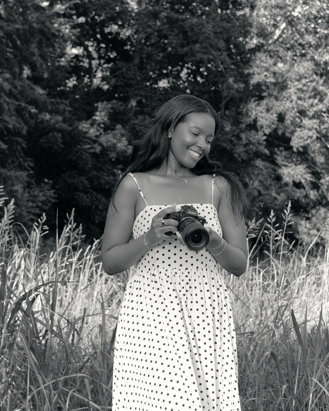 Smiling woman in polka dot dress holding a camera in a black and white outdoor portrait.
