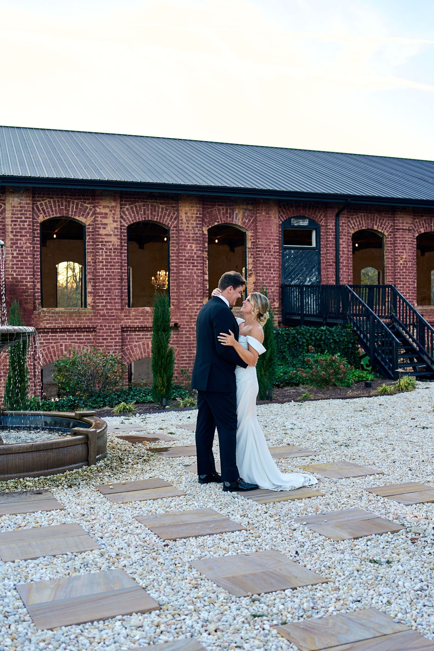A couple shares a romantic moment on a stone patio in front of a historic red brick building with arched windows.