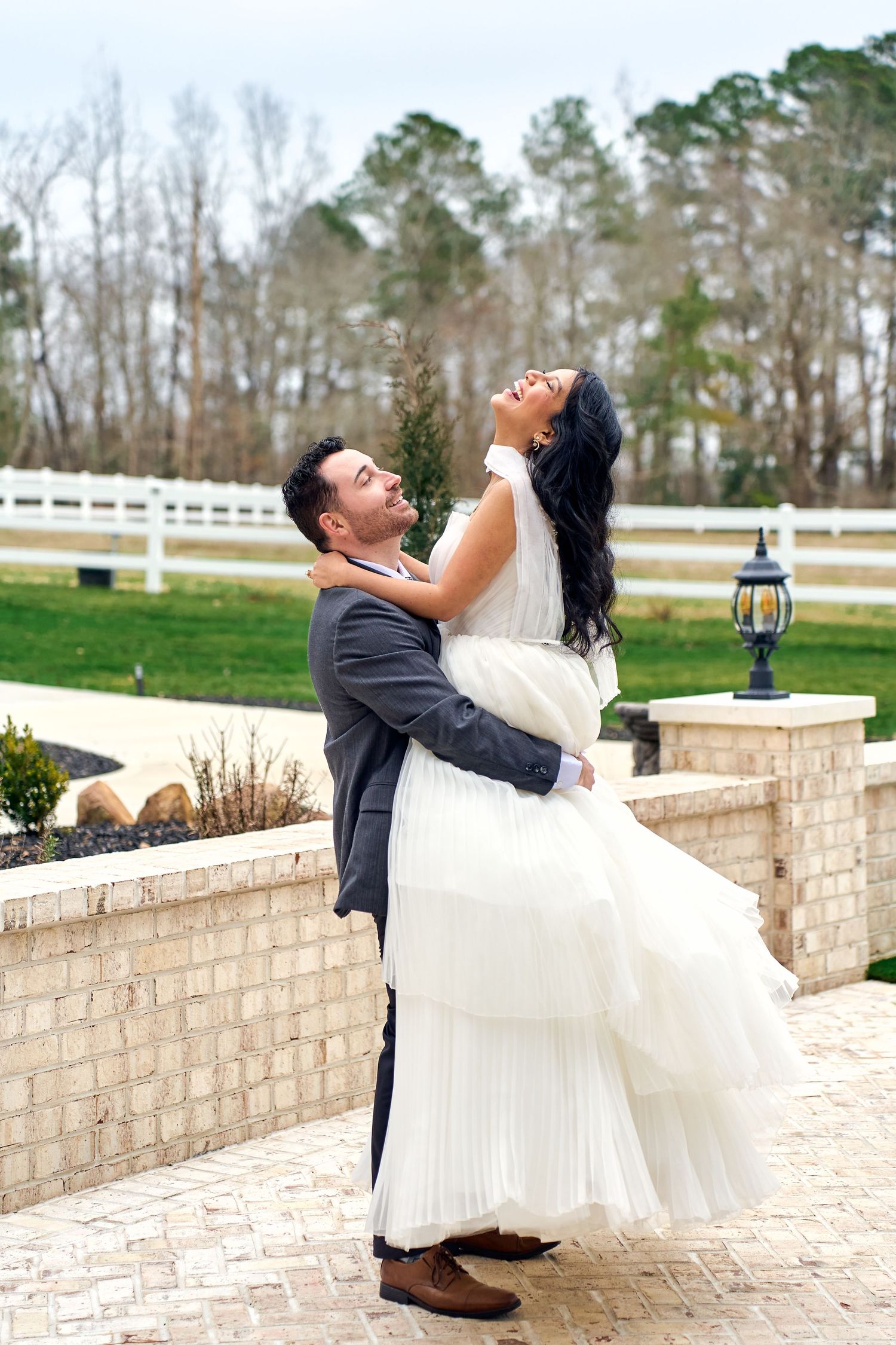 Happy newlyweds share a joyful embrace on brick patio with white fence and rustic lantern in countryside setting.