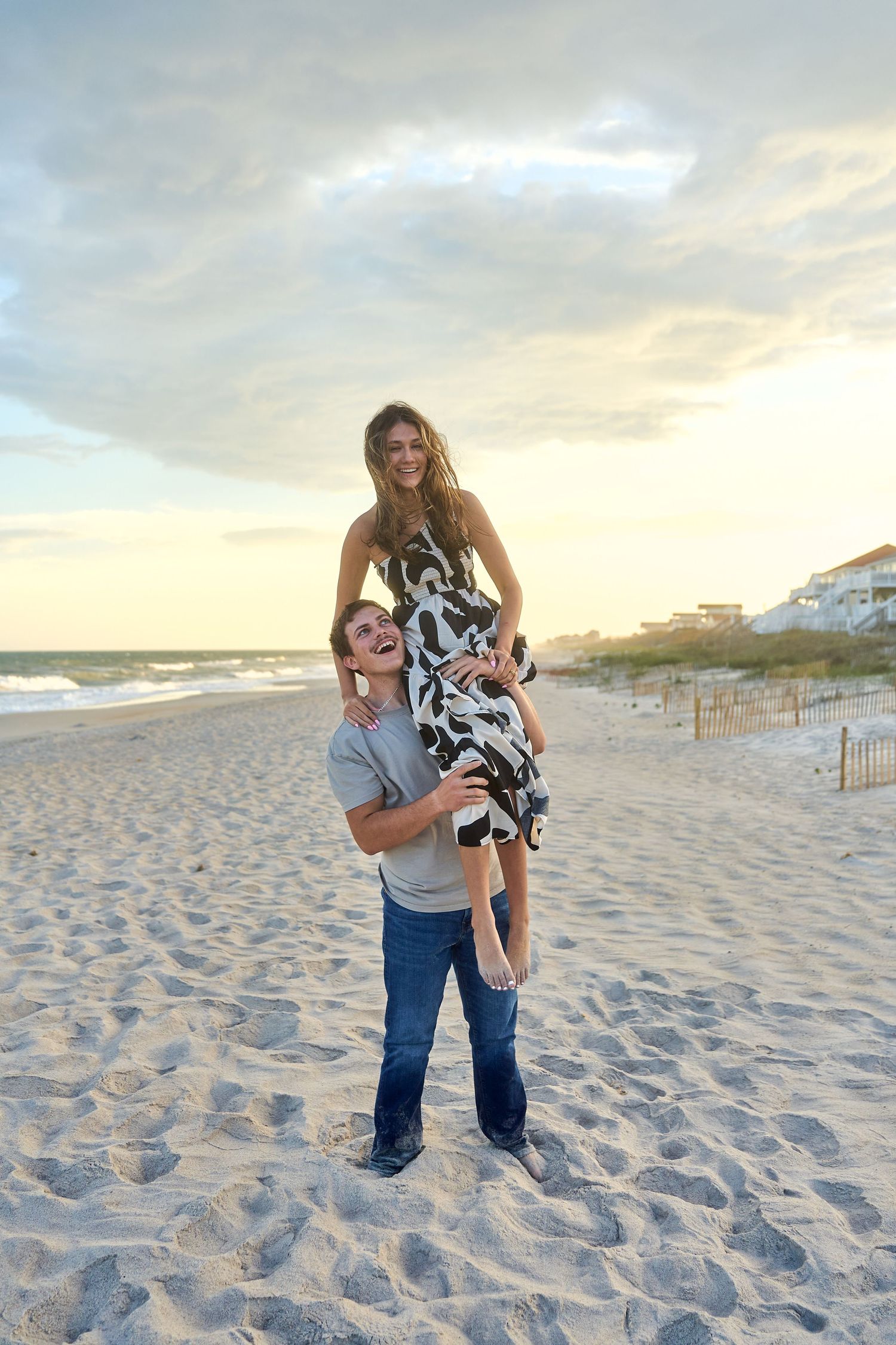 A couple shares a playful moment on a sandy beach at sunset with coastal homes visible in the background.