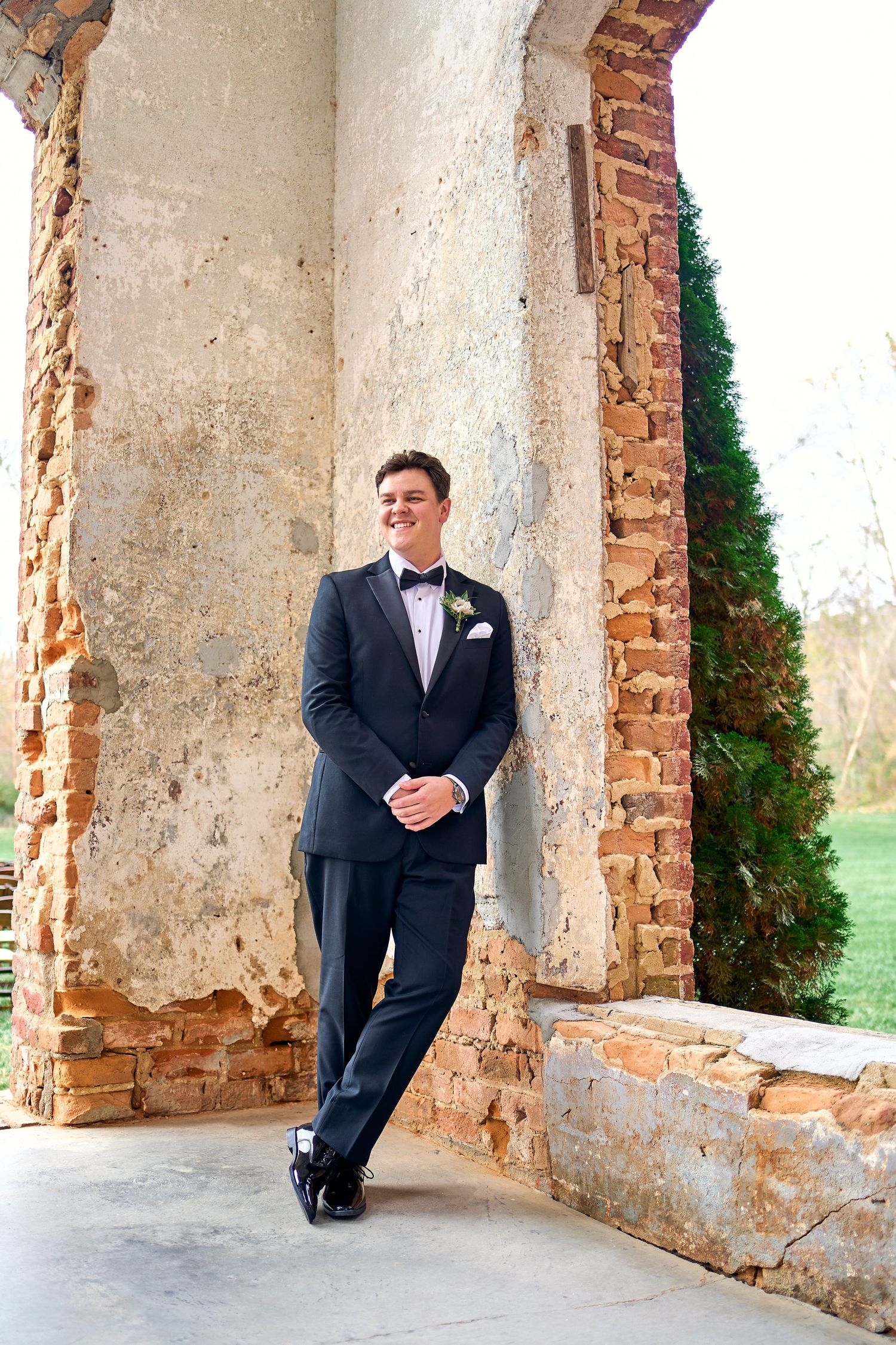 A person in a dark suit and bow tie leans against an old stone archway with exposed brick and plaster walls.