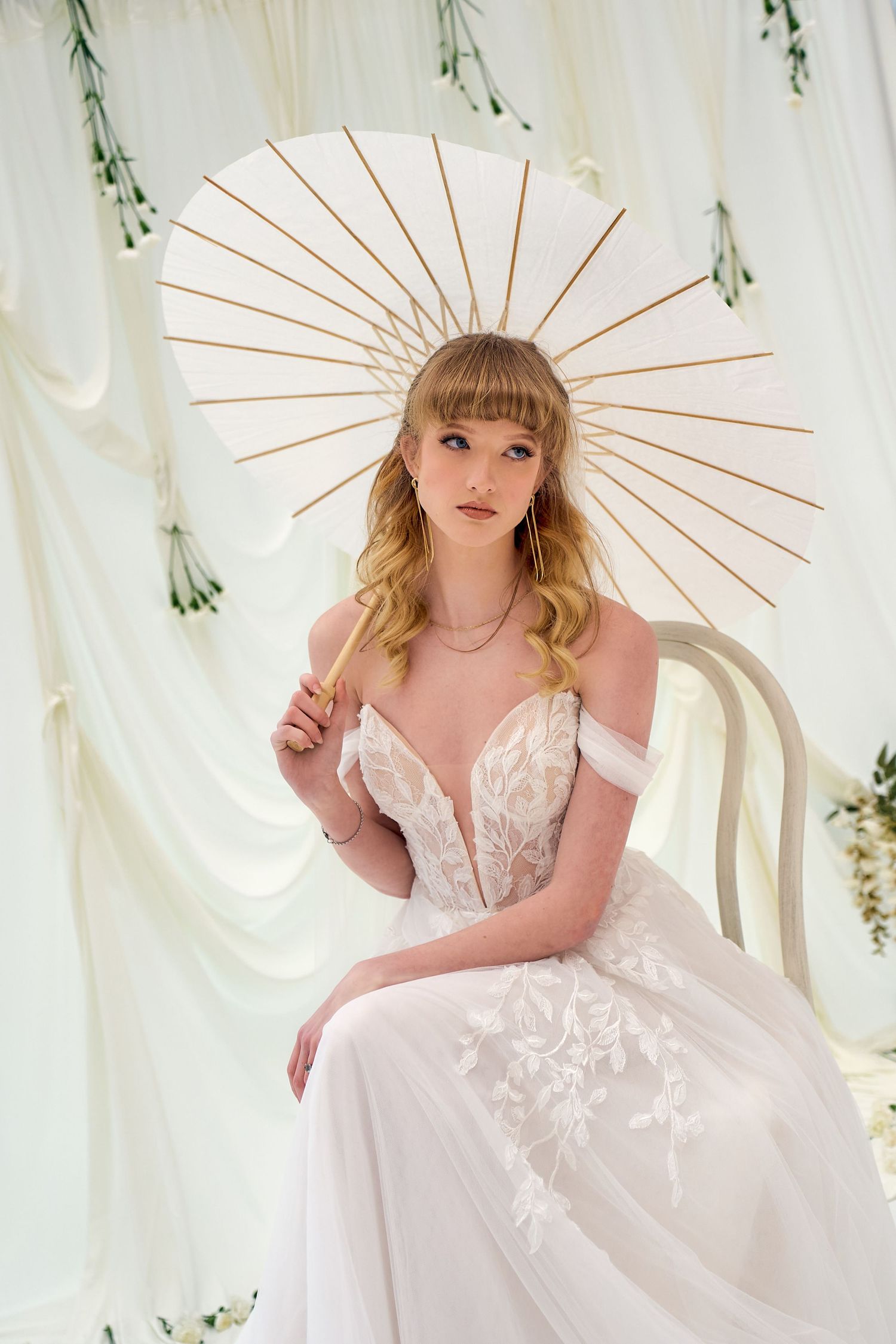 Wedding dress with plunging neckline and floral details shown with decorative white parasol against draped fabric backdrop.