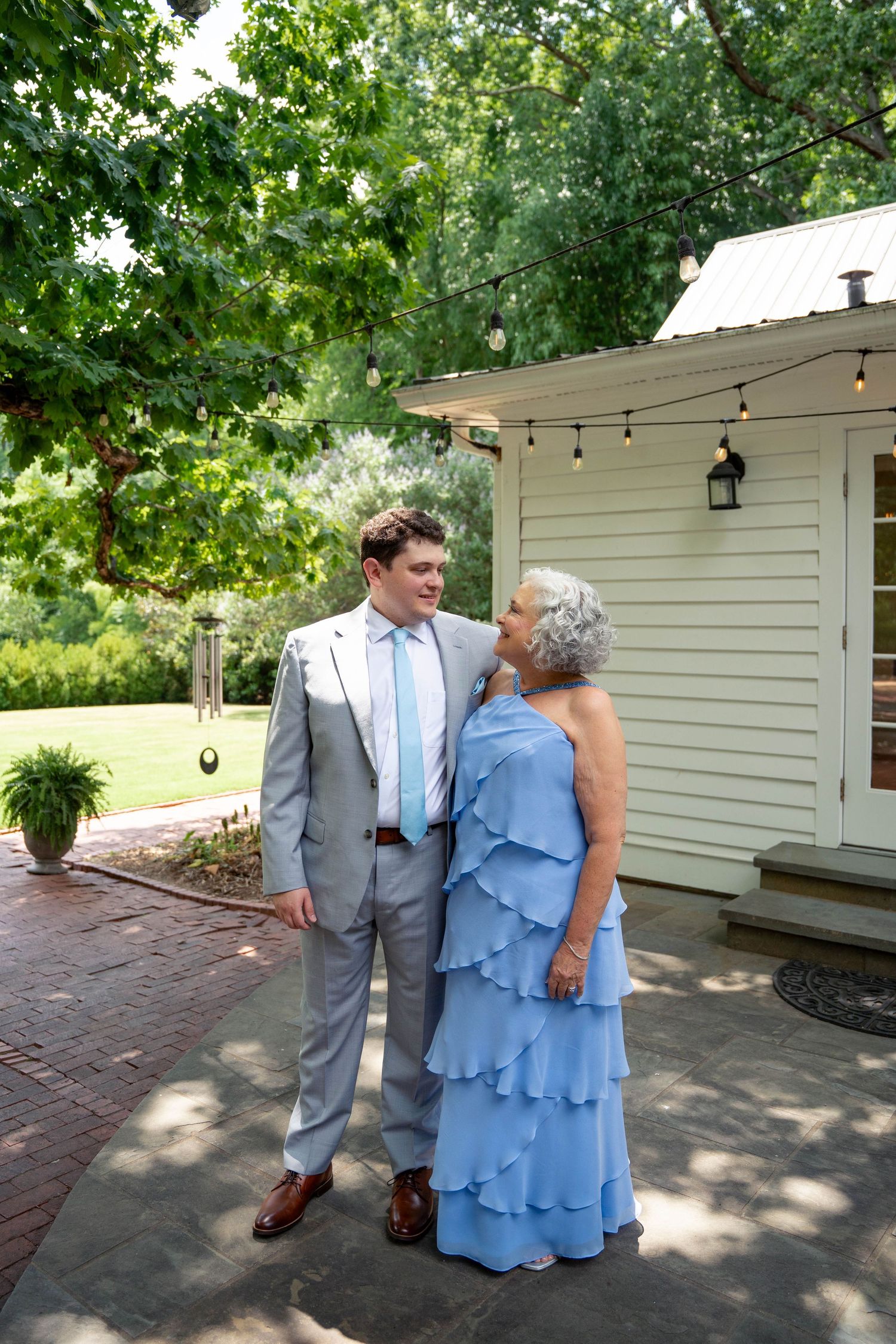 Groom in gray suit shares a tender moment with elderly woman in blue ruffled dress at wedding venue.