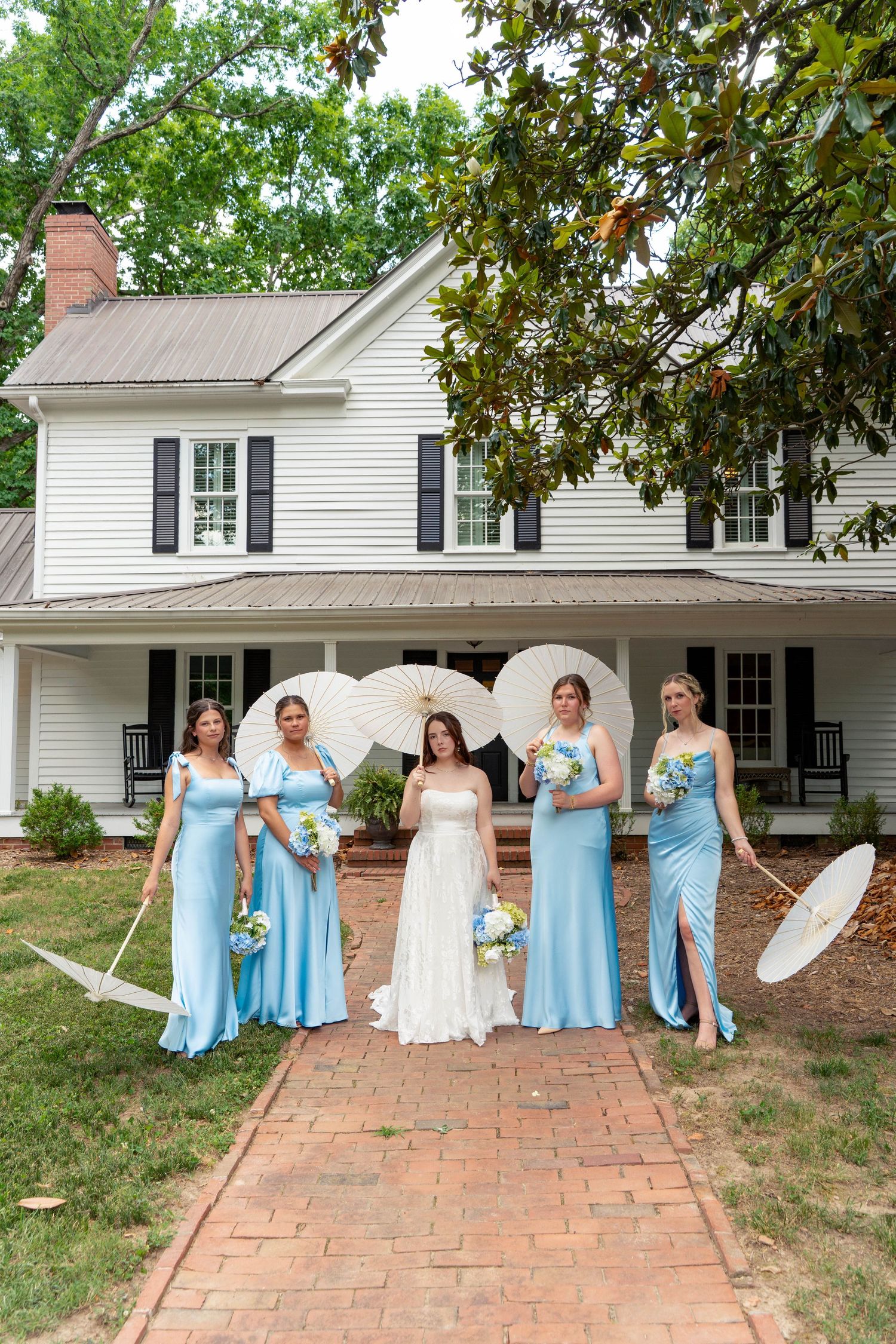 Bride with bridesmaids in blue gowns holding white parasols and bouquets outside white house.