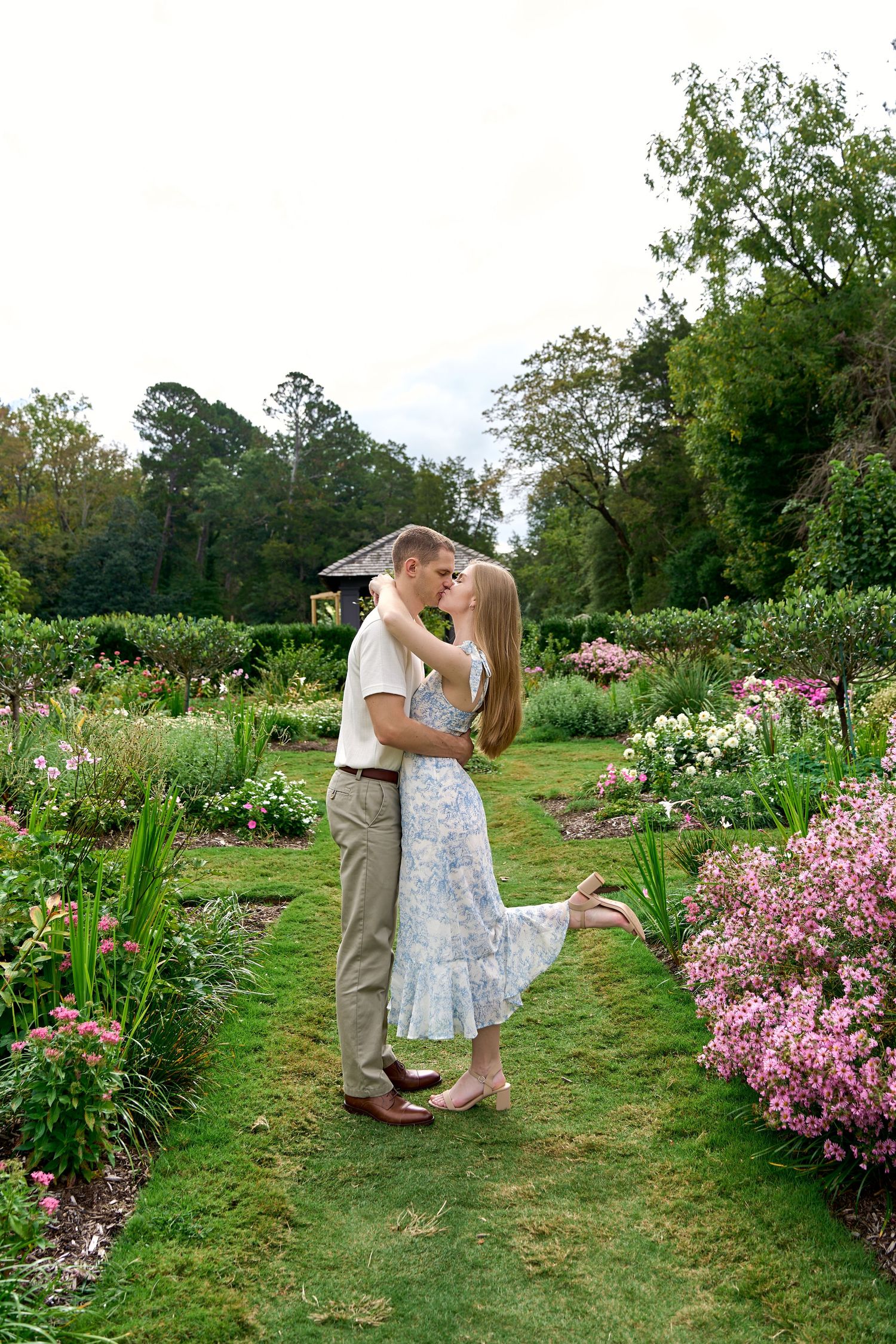Couple sharing a romantic kiss surrounded by colorful blooming flowers in a lush garden pathway.
