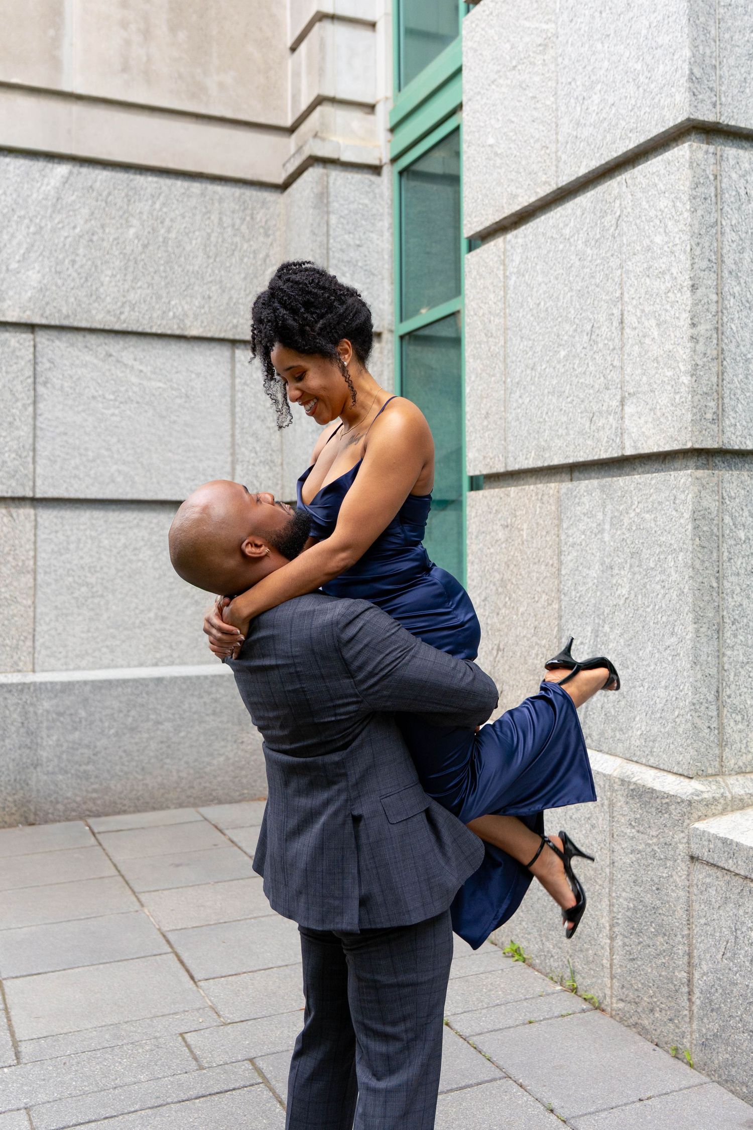 Joyful Black man lifting smiling woman in navy satin dress against a stone building exterior.
