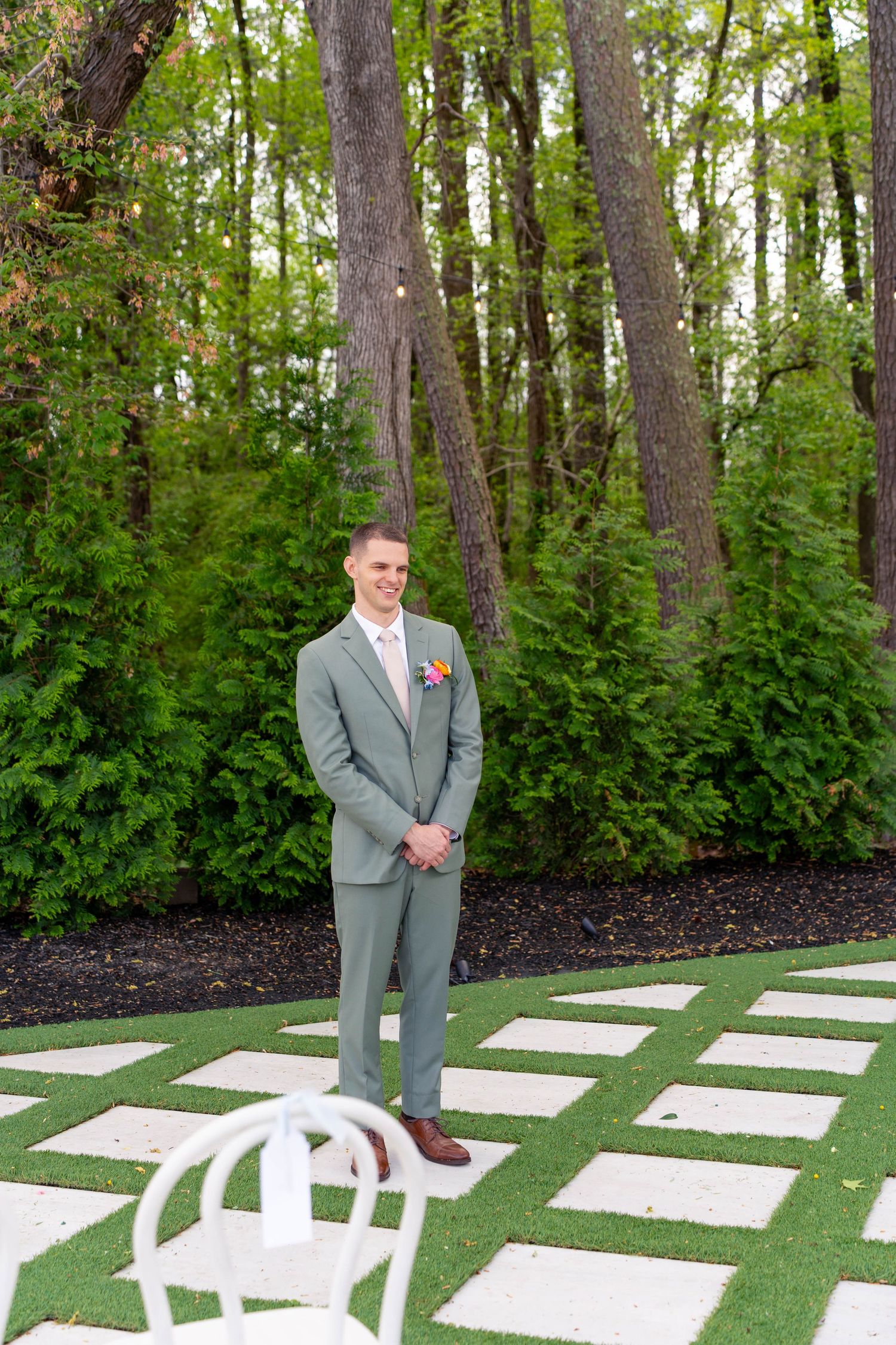 Groom in sage green suit stands confidently on a stone pathway surrounded by lush green trees.
