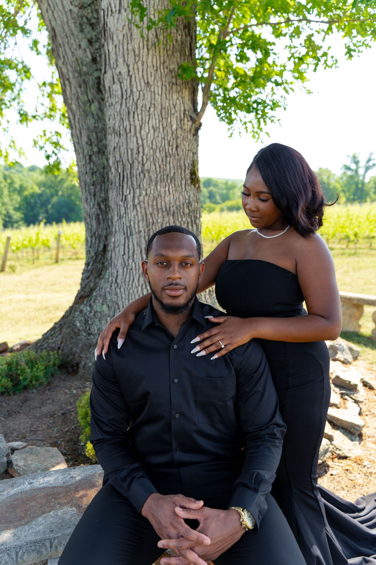 Couple posing under large tree in vineyard, man seated on stone bench with woman behind him.