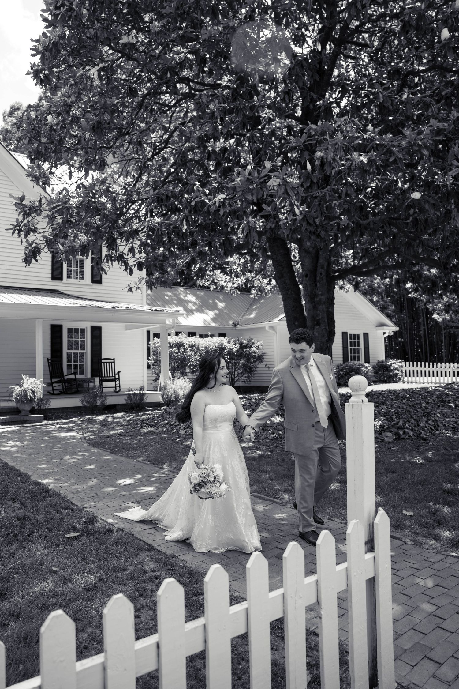 Black and white photo of bride and groom walking by white picket fence.