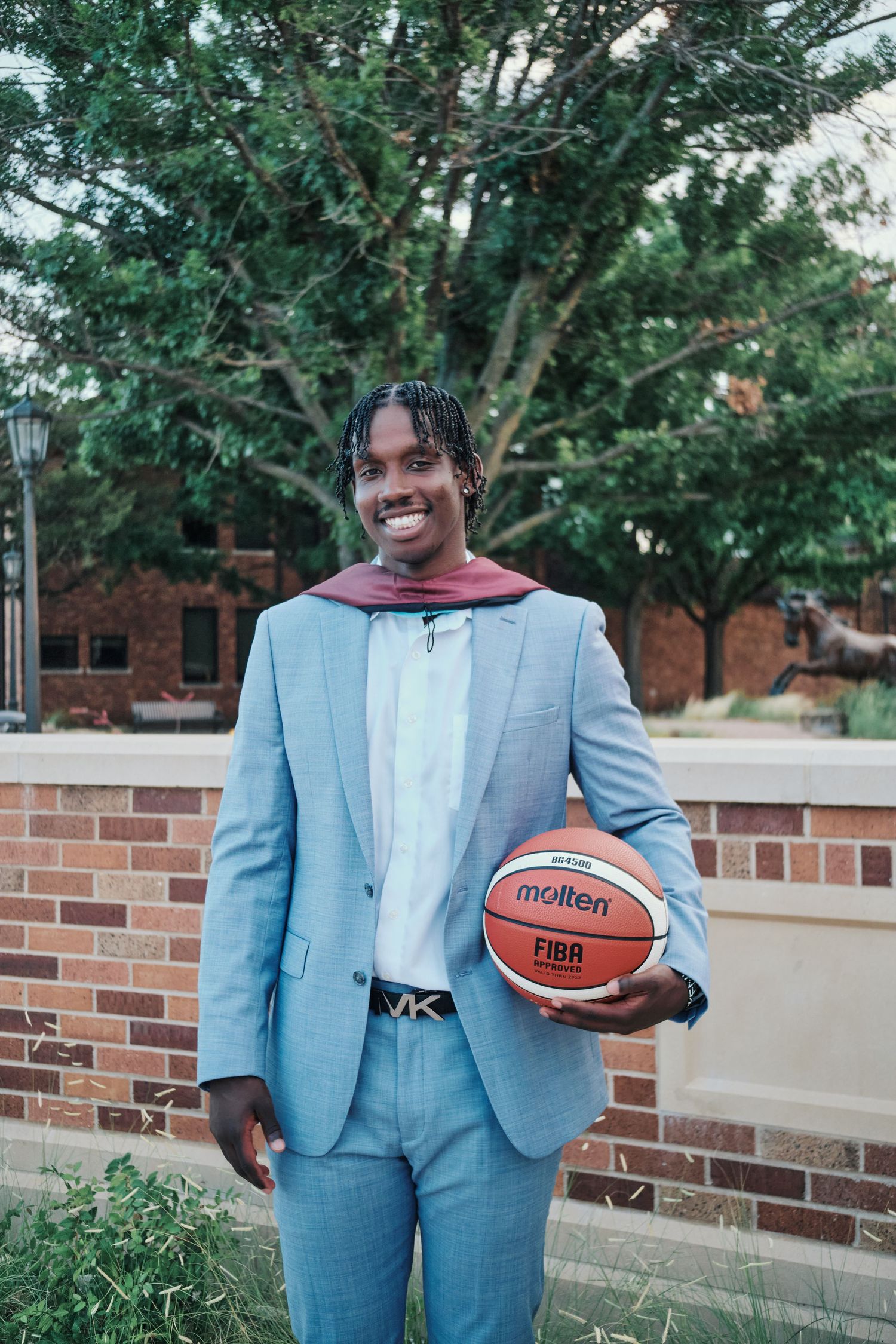 Young man in light blue suit holding a Molten FIBA basketball smiling outdoors near brick wall.