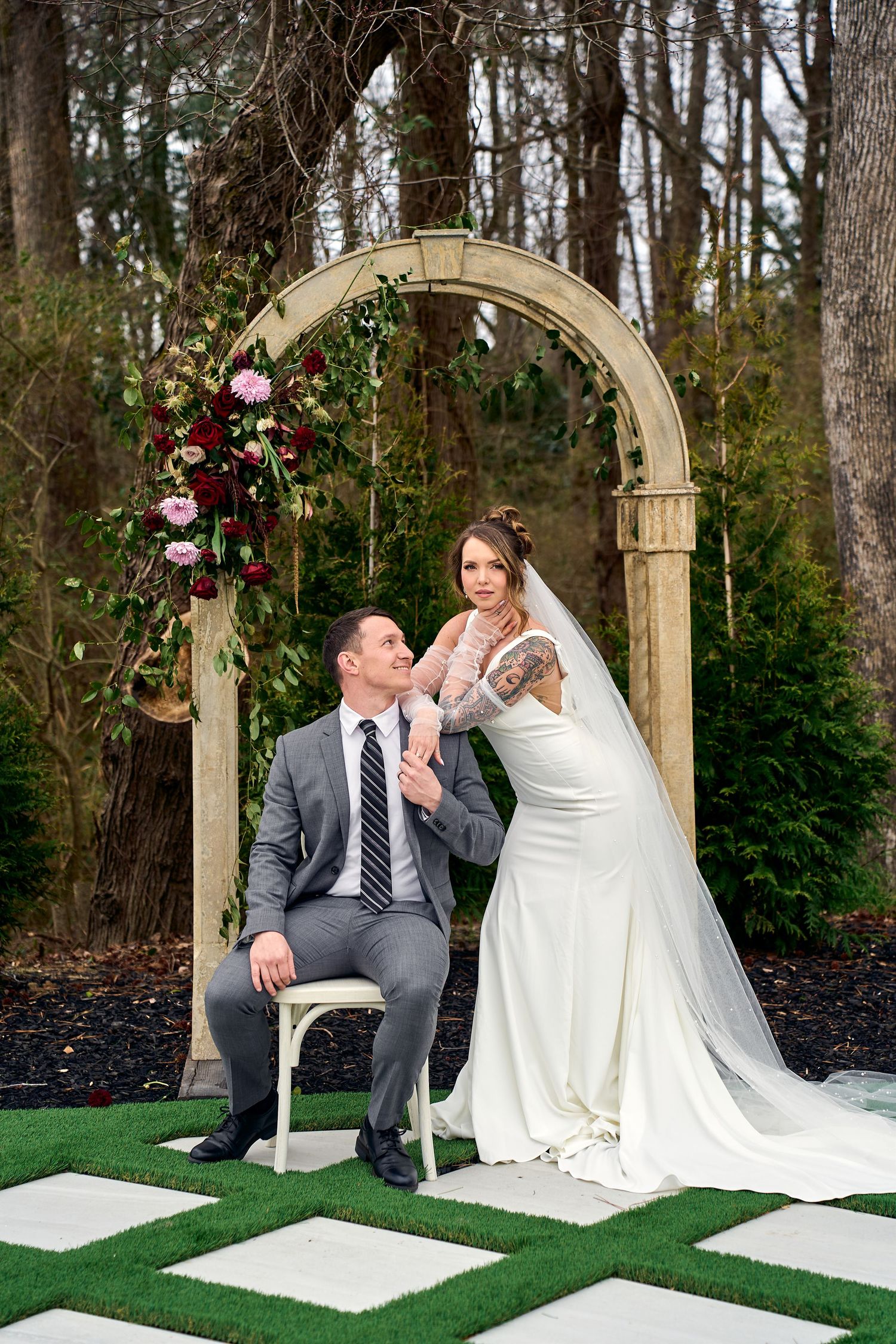 Romantic outdoor wedding ceremony setup with an arch decorated with burgundy and pink flowers on a paved garden path.