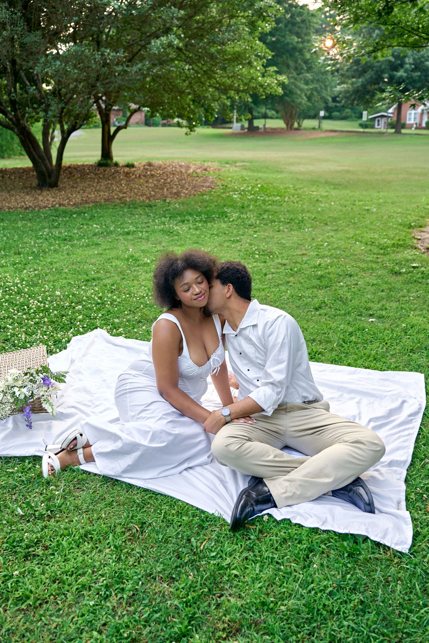 A romantic picnic scene on green grass with a white blanket and flowers in a peaceful park setting.