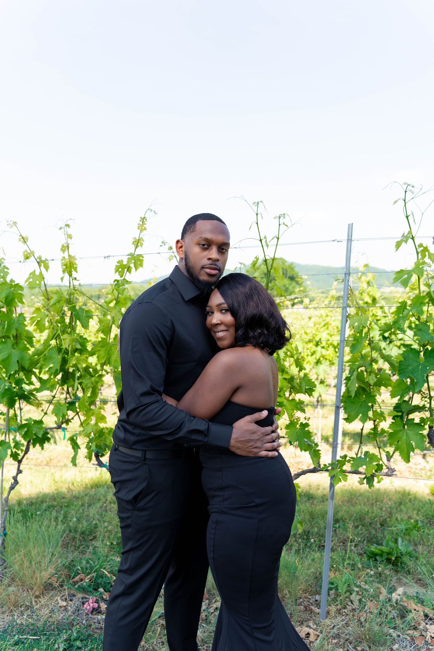Couple in matching black outfits embracing warmly in front of lush green vineyard rows at dusk.