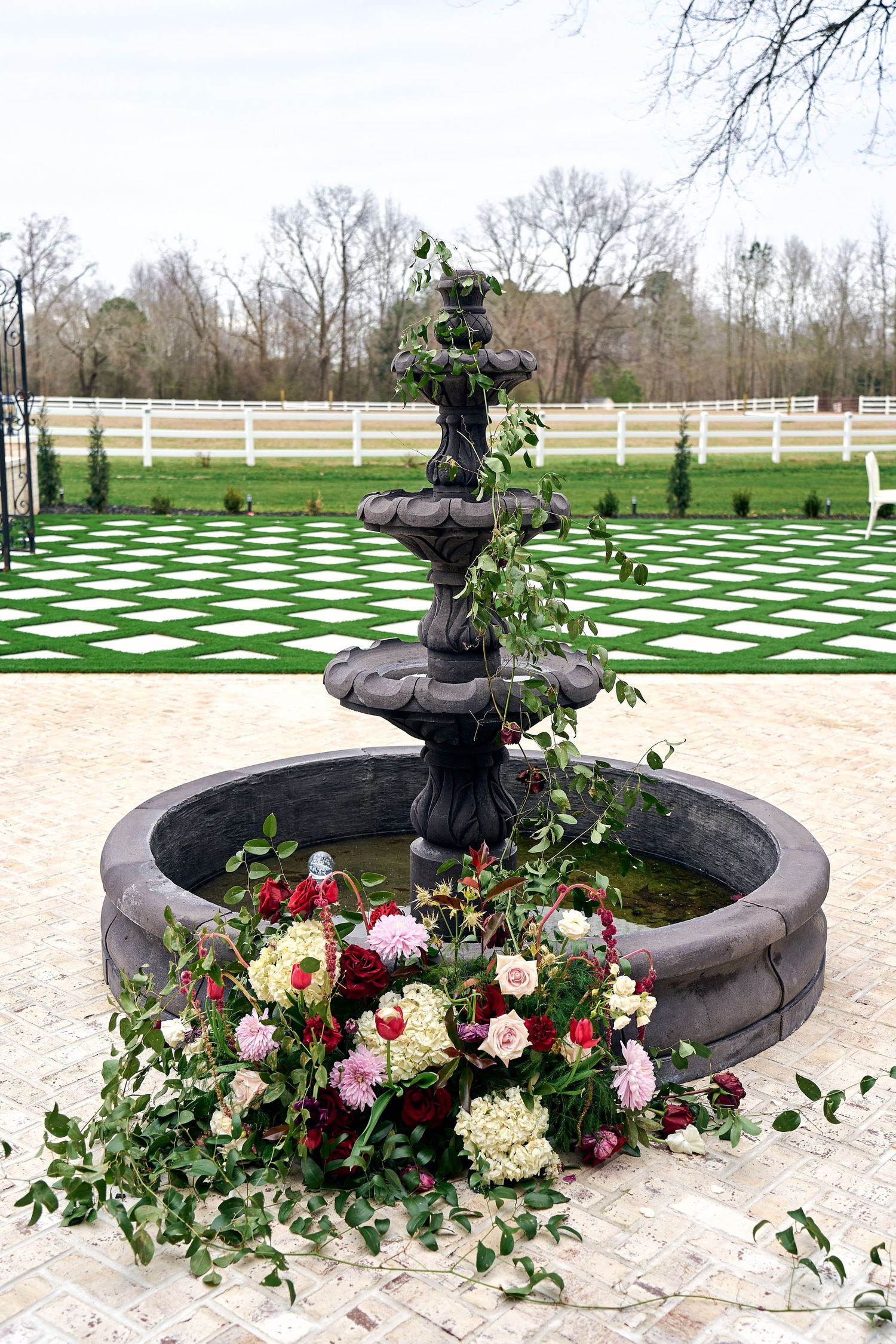 A decorative garden fountain with cascading tiers adorned with pink and red roses and trailing vines on a checkerboard patio.