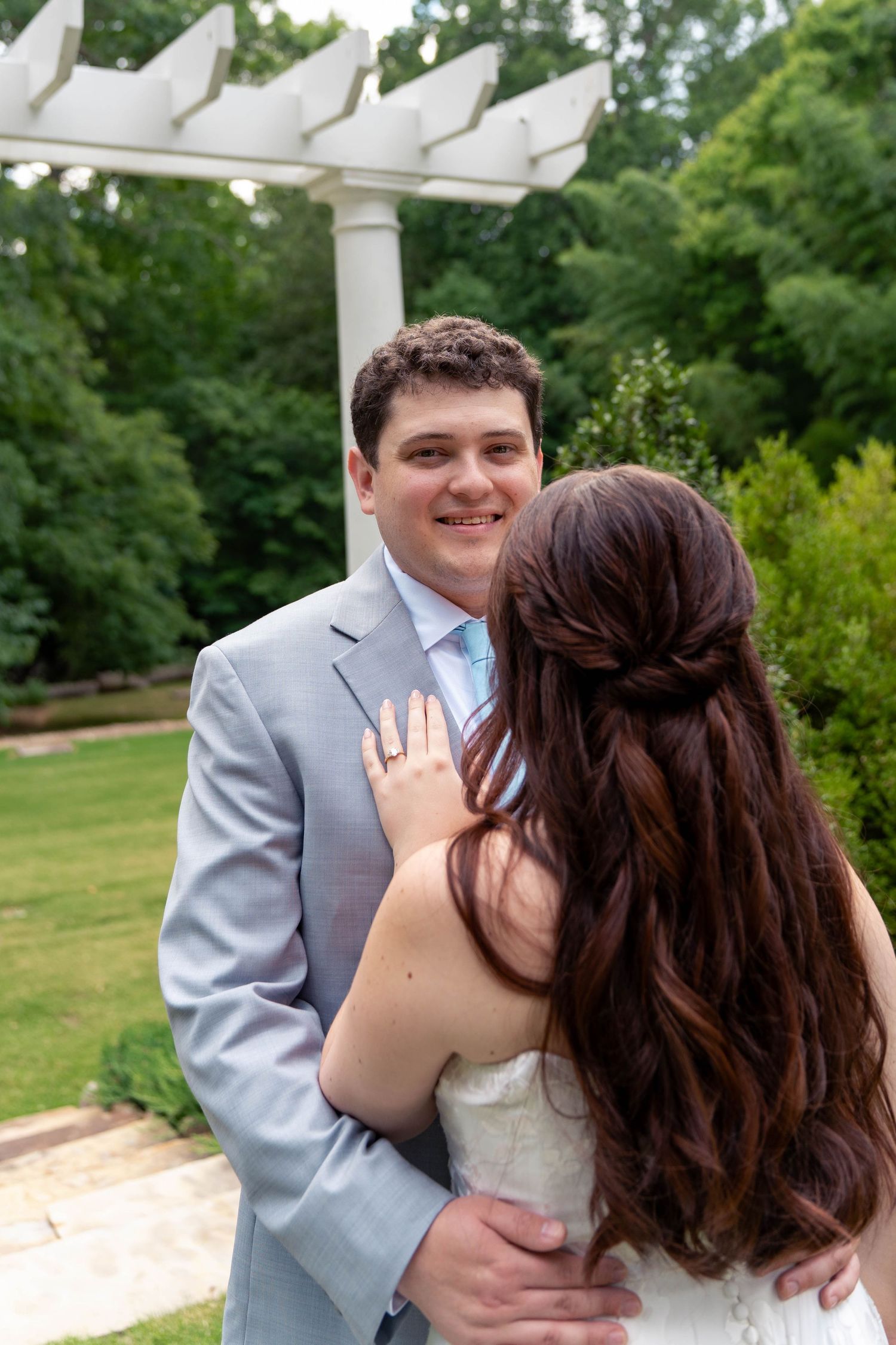 Smiling groom in gray suit embracing bride in lush green garden setting.
