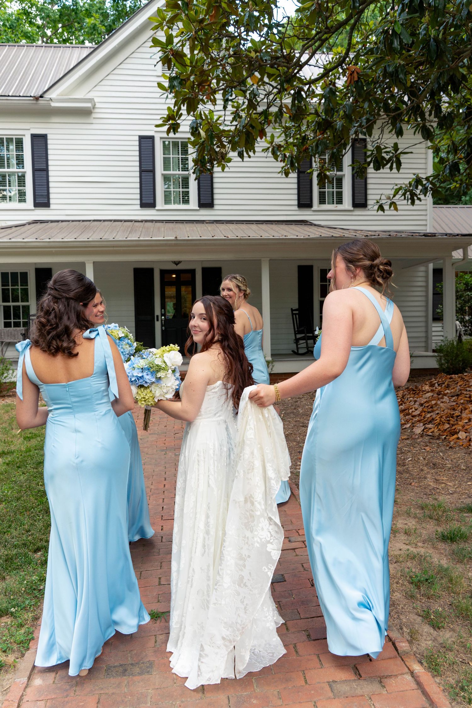 Bride in white lace gown walks with bridesmaids in blue dresses on brick path toward white house.