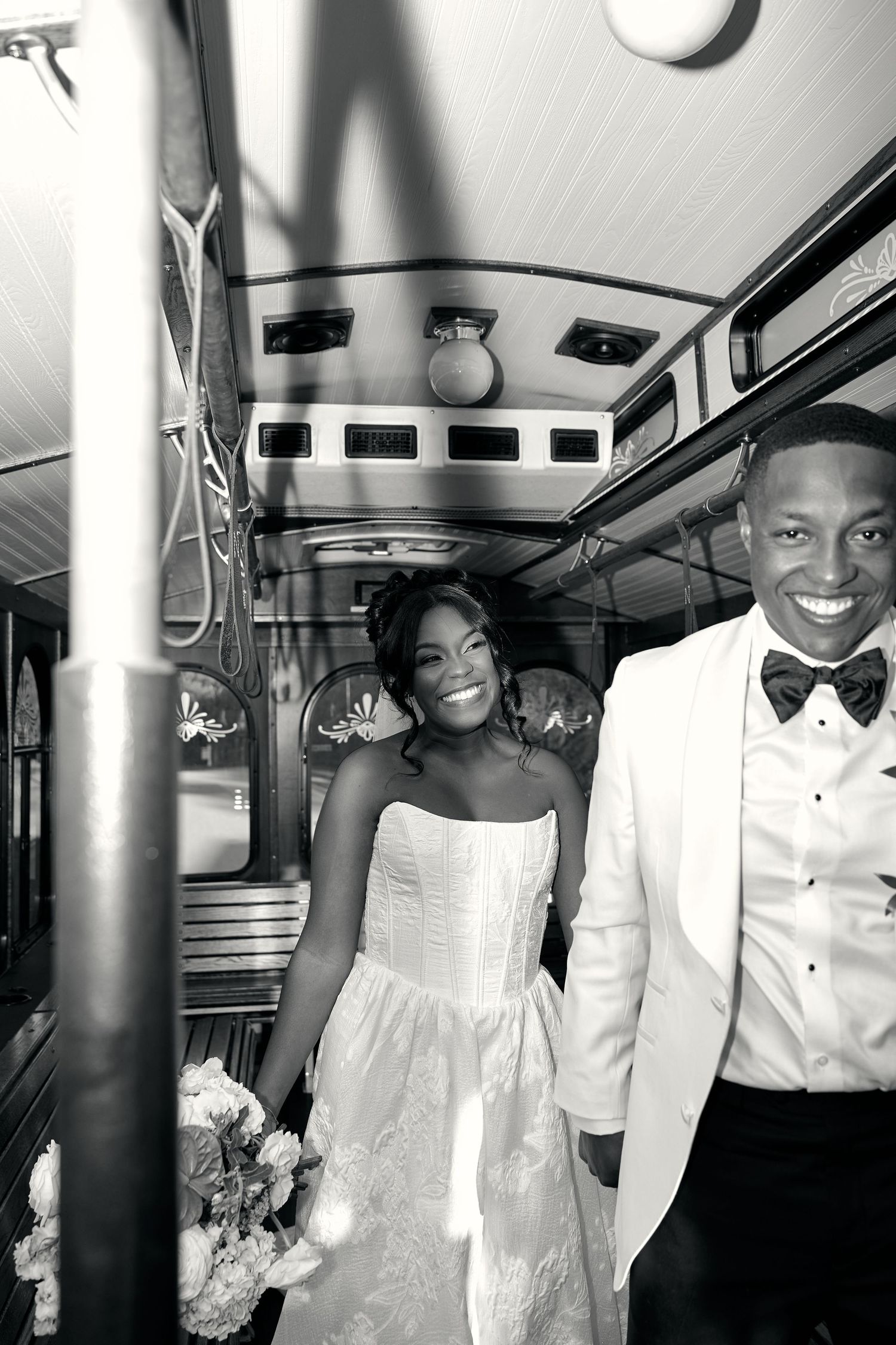 Black and white photo of smiling bride and groom riding together on a vintage trolley bus.