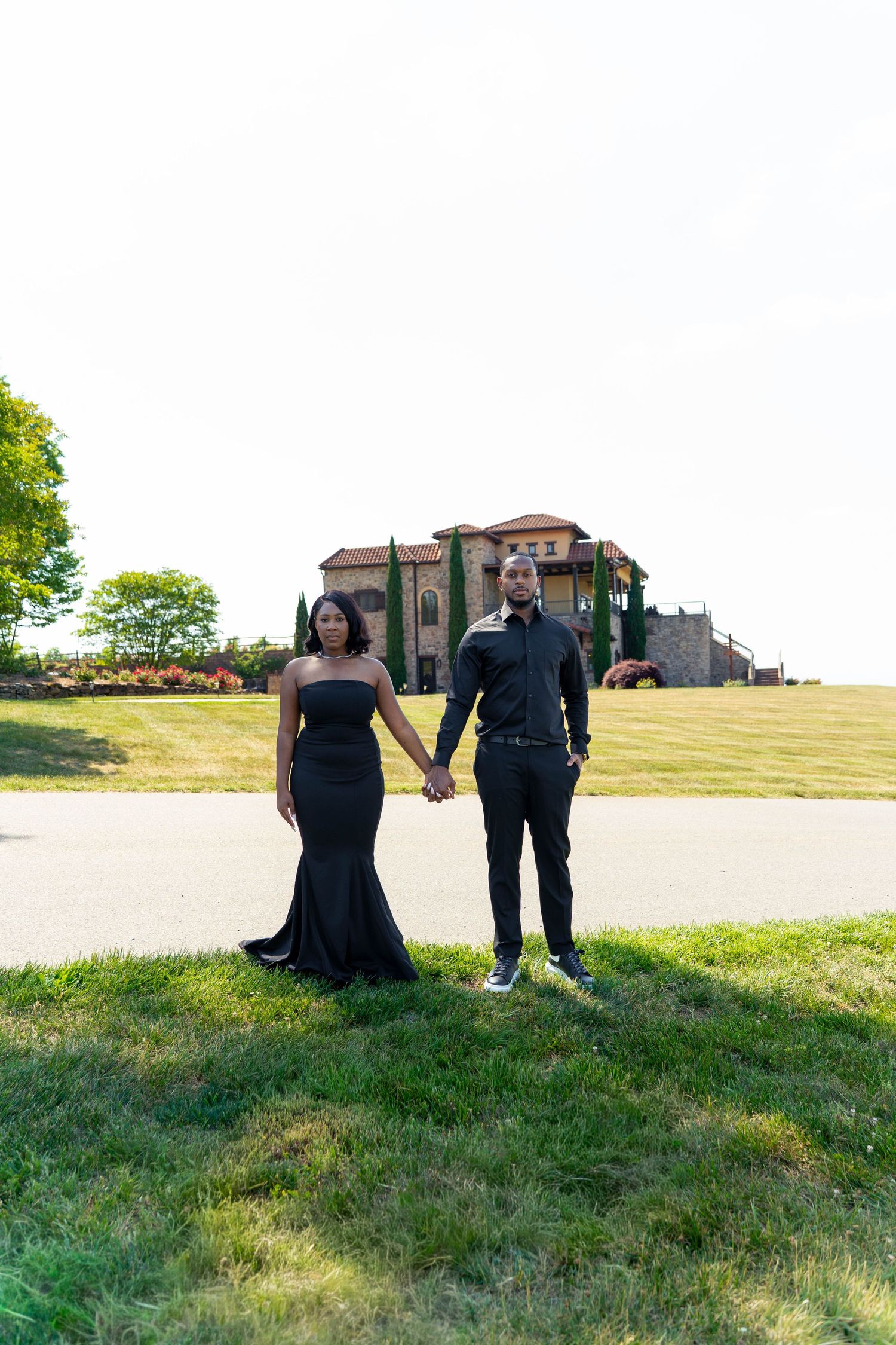 Elegant couple in matching black outfits holding hands on green lawn with Italian-style mansion in background.